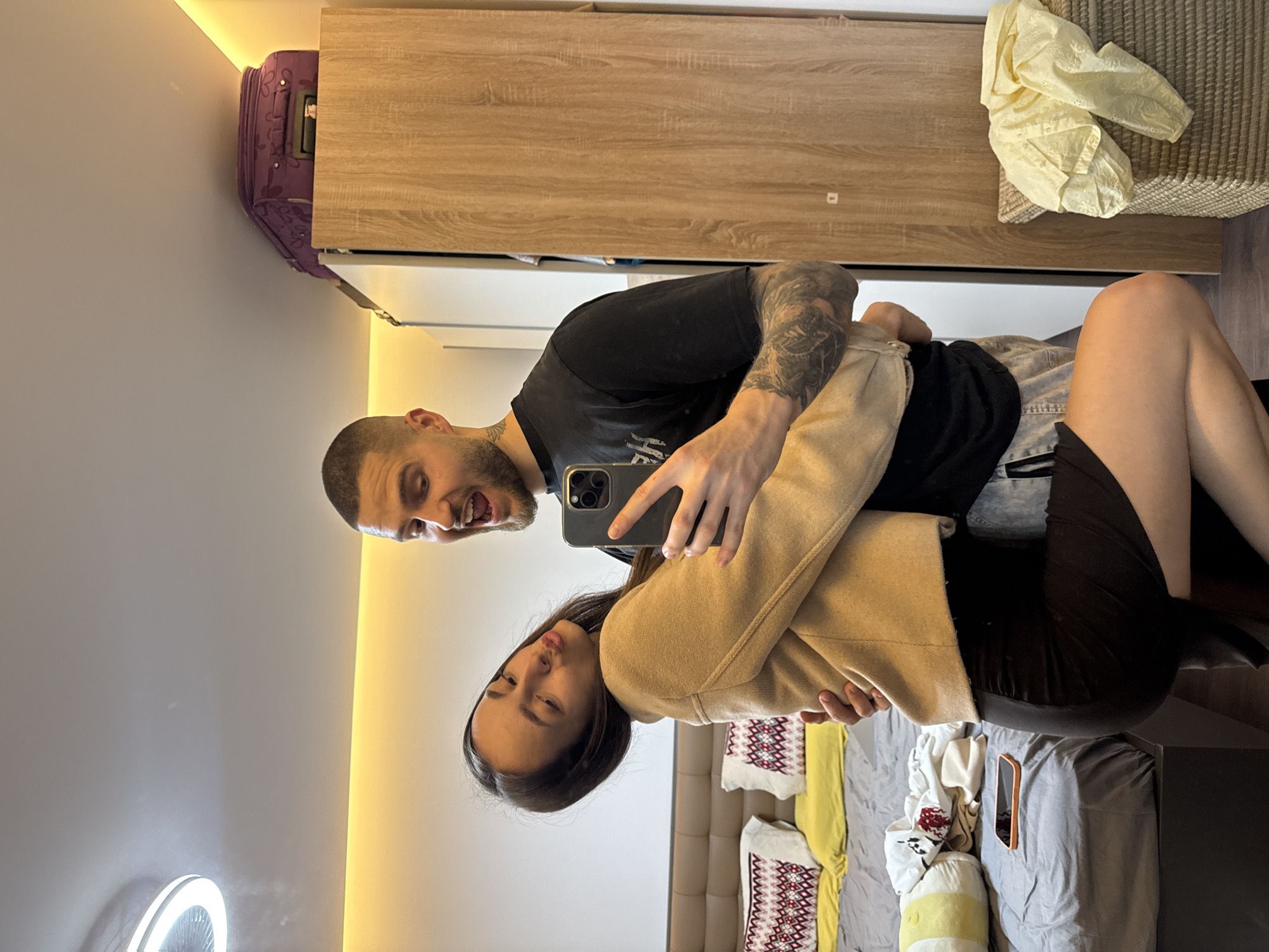 A man and woman are posing for a selfie in a room. The man is wearing a black t-shirt with arm tattoos. He is holding a smartphone. The woman is wearing a light brown coat. The room contains a bed with pillows and linens, a wardrobe, a suitcase, a round mirror with a light, and a wall with a light fixture. The specific location in Sofia, Bulgaria is not discernible from the image.