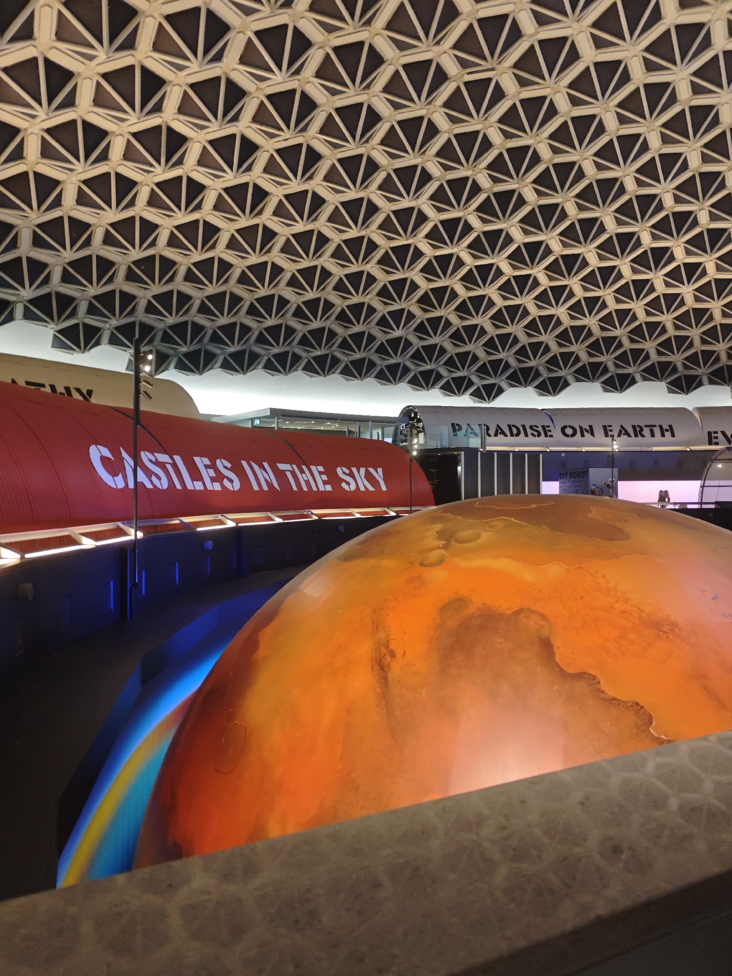 The image captures an indoor exhibition space, likely a museum or science center, characterized by its unique architectural ceiling composed of interconnected triangular panels. In the foreground, a large, rounded object with an orange and reddish hue dominates, possibly a model of a planet, with a luminous blue and yellow band visible to its left. Behind this, a large red cylindrical structure displays white text that reads "CASTLES IN THE SKY." Further back, another elongated structure, appearing white and metallic, bears the text "PARADISE ON EARTH" in black lettering. The lighting in the space is artificial, with visible light fixtures illuminating the exhibits. The overall impression is one of a themed exhibition, possibly related to space, science, or imagination, set within a modern, geometrically designed interior. The location is identified as Eindhoven, Netherlands.