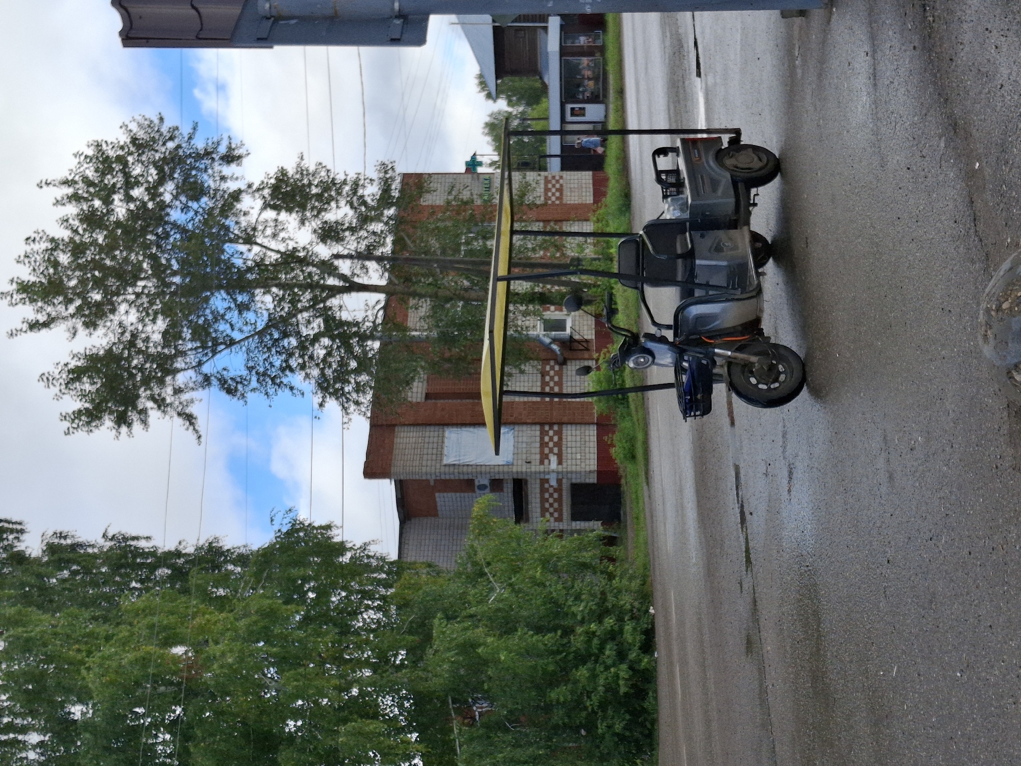 In Elovo, Russia, a three-wheeled electric scooter with a yellow canopy is parked on a wet asphalt road. The scooter has two seats, the front seat for a driver and a rear seat for a passenger. The scooter is facing towards the right of the image. Behind the scooter, there is a brick building with a white and red facade, and a small kiosk-like structure with a glass front. Trees and power lines are visible in the background, under a cloudy sky. The overall scene depicts a quiet street in what appears to be a residential or commercial area.