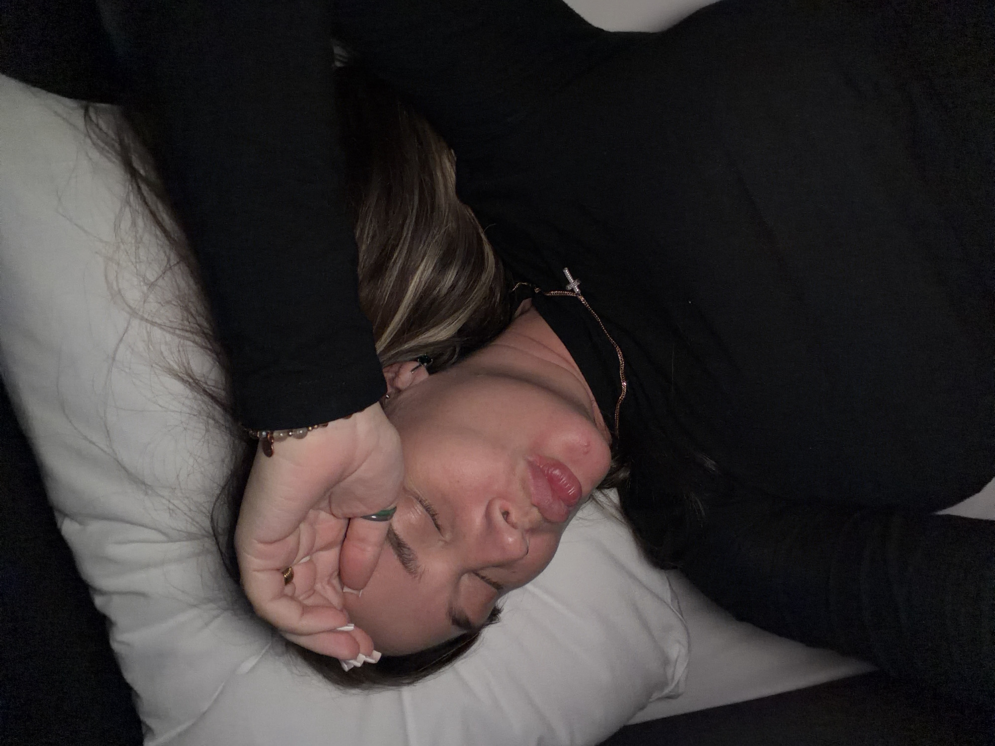 A woman is lying on a pillow, her eyes closed and her hand resting on her forehead. She is wearing a black long-sleeved shirt and a necklace. Her hair is long and dark with blonde highlights. The background is a pillow and a dark surface. There are no discernible elements in the image that would indicate a location in the city of Solov'evo, Russia.