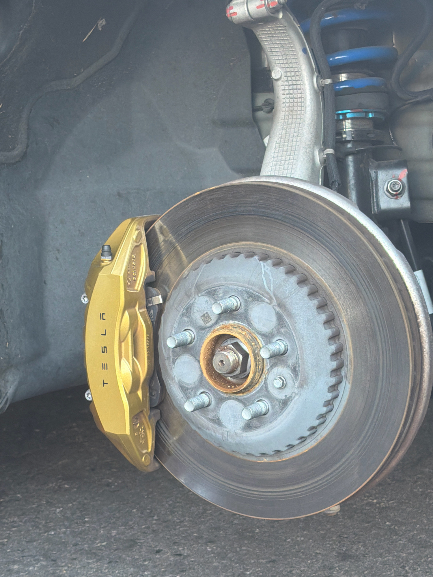 The image shows a close-up view of a car's braking system, specifically the front passenger-side wheel well of a Tesla. The caliper, painted a bright gold, prominently displays the "TESLA" logo. The brake rotor is visible behind the caliper, showing signs of wear. The suspension components, including a blue coil spring and a silver control arm, are also visible. The scene appears to be outdoors, with asphalt visible in the foreground and a dark, textured surface suggesting the wheel well liner. The overall lighting suggests it is daytime. No people are present in the image. The location is not specified in the image, and therefore cannot be determined to be in Los Angeles, California, United States of America.