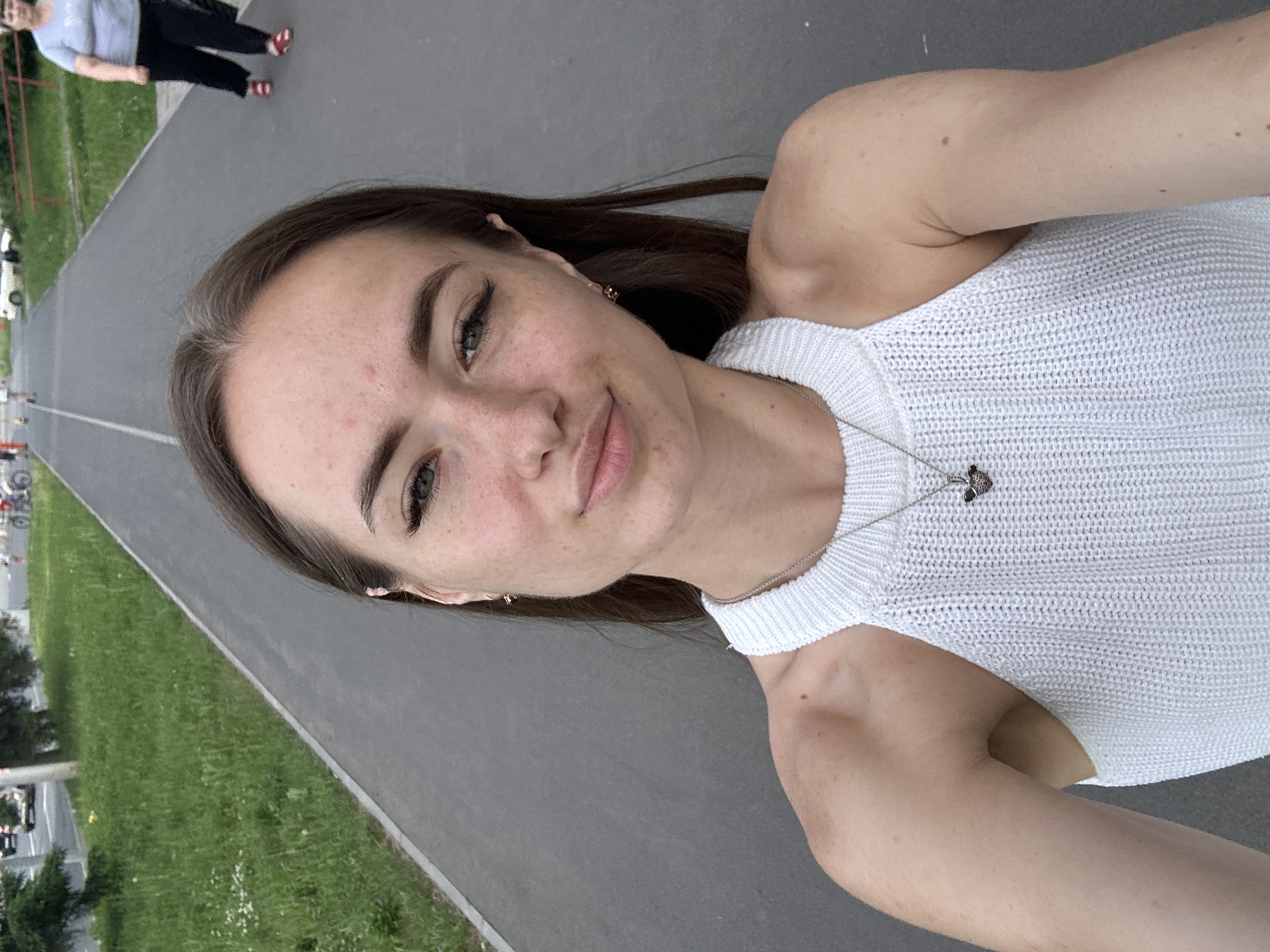 An outdoor, medium shot captures a young woman with long brown hair and freckles on her face, posing for a selfie. She is wearing a white knitted halter top and a silver necklace with a heart pendant. Her eyes are green, and she has on eyeliner and mascara. In the background, a paved path runs alongside a grassy area. To the right of the path, a woman in a light blue t-shirt and black pants walks away from the camera, wearing red sandals. Further in the distance, other figures and vehicles are visible. The scene appears to be a public space within a city.