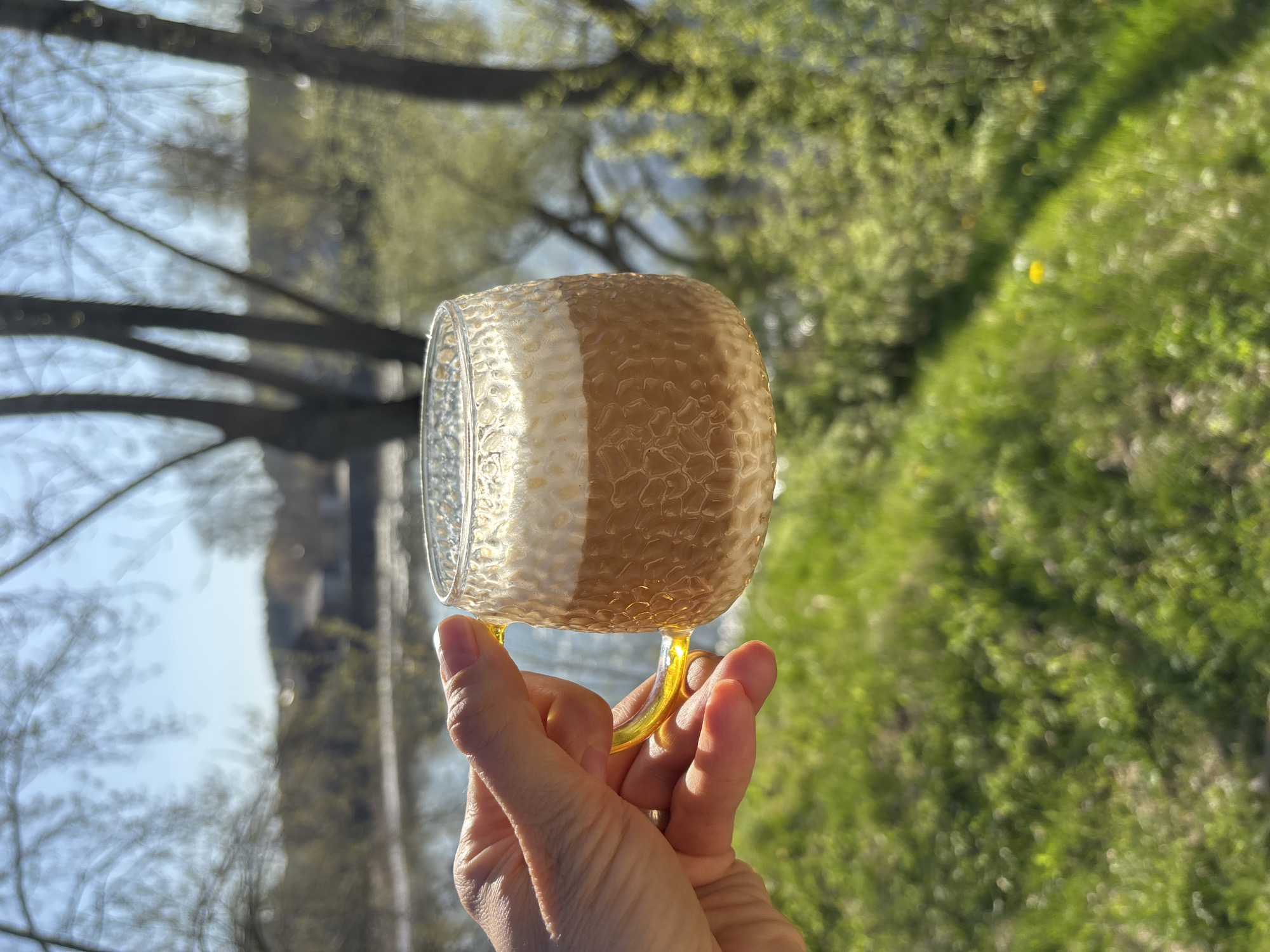 A close-up image shows a hand holding a glass mug filled with a beverage, likely coffee with milk, outdoors. The mug has a textured, honeycomb-like design and a yellow handle. The background is blurred but shows green foliage, suggesting a park or garden setting, and what seems to be a building reflected in the glass. The image orientation is vertical.
While the city of Chaikovsky, Russia is requested, there is no contextual data within the image to confirm that location.