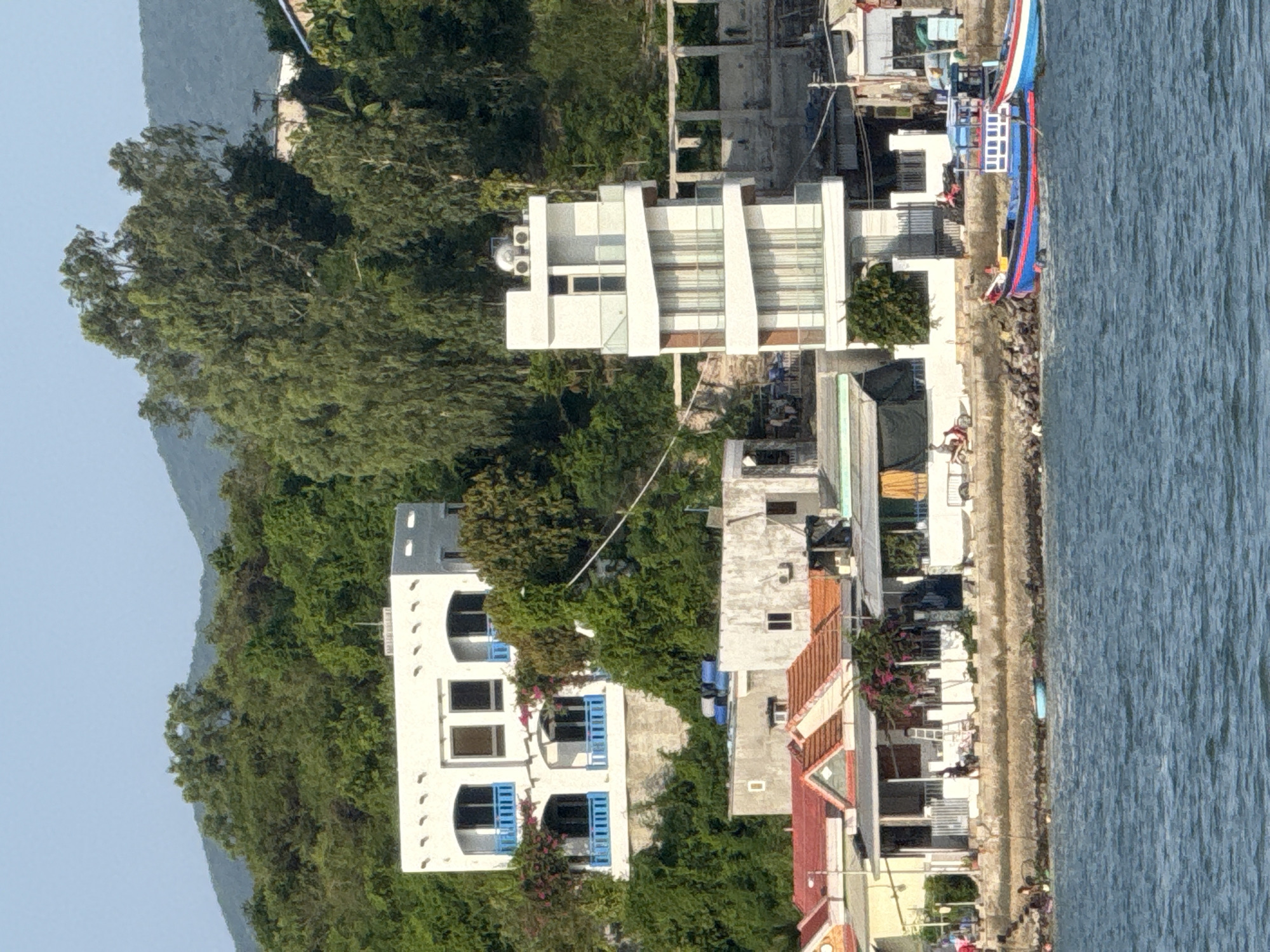 The image depicts a waterfront scene in Nha Trang Ward, Vietnam. In the foreground, the blue-green water of the sea is visible with moderate ripples. Two boats are docked along the shore. The larger boat is predominantly blue with red and white accents, featuring a white structure on deck with windows. The smaller boat, to its right, is partially visible and also has red and white colors.
The shoreline is rocky and lined with a low stone wall. Behind the wall, a street or walkway runs parallel to the water. Along this walkway are several buildings and structures. Closest to the water are single-story houses with red-tiled roofs. Further back, a more modern, multi-story white building with a distinctive, staggered design stands out. It features balconies with glass railings and large windows. To the left of this modern building, a larger, white, two-story house with blue balconies and decorative trim is situated on a hillside. This house is nestled amongst lush green trees and vegetation, as are the other buildings in the background, which ascend a green, tree-covered hill.
Several people are visible near the buildings, including individuals walking along the walkway and one person riding a red scooter. Laundry is hanging on some of the buildings. The sky is clear and pale blue. The overall impression is a coastal town with a mix of traditional and modern architecture, surrounded by abundant greenery.