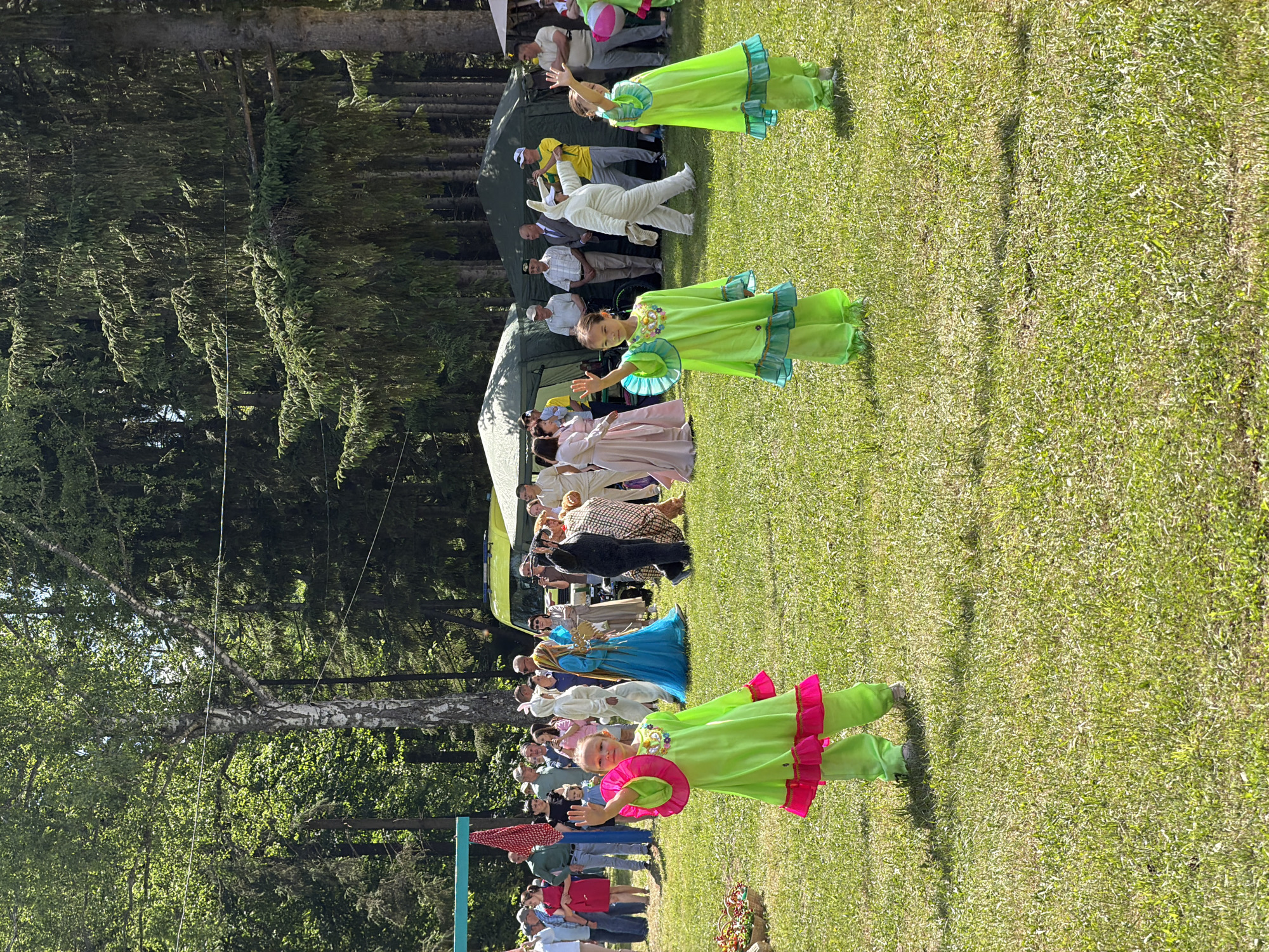 The image depicts an outdoor event taking place on a well-maintained grassy field under a bright, clear sky. In the immediate foreground, three young girls, approximately 5-10 years old, are positioned as if performing or posing. The girl on the far left wears a lime green two-piece outfit with bright pink ruffled trim on the sleeves and hem, and is smiling directly at the viewer while raising her right hand in a wave. The girl to her right (center-left) wears a similar lime green outfit with teal ruffled trim, and is also facing forward, raising both hands in a wave. The third girl, partially visible on the far right, also wears a lime green and teal outfit and appears to be waving.
Behind the girls, a diverse crowd of people is gathered. Several individuals are in elaborate costumes, including a person in a full white rabbit suit, a person in a long blue gown with a golden headpiece, and two individuals in dark and patterned animal or character suits. Other people in the crowd are dressed in casual attire, observing the scene, with some holding mobile phones.
In the midground, two large, olive-green canvas tents, possibly military-style, are erected. A yellow emergency vehicle, resembling an ambulance, is partially visible behind the tents on the right side. The background consists of a dense forest with a mix of tall coniferous trees and a prominent birch tree with white bark. The sunlight is bright, casting distinct shadows on the grass. The event appears to be a public gathering, possibly a festival or performance, in an outdoor setting in Unknown, Unknown.