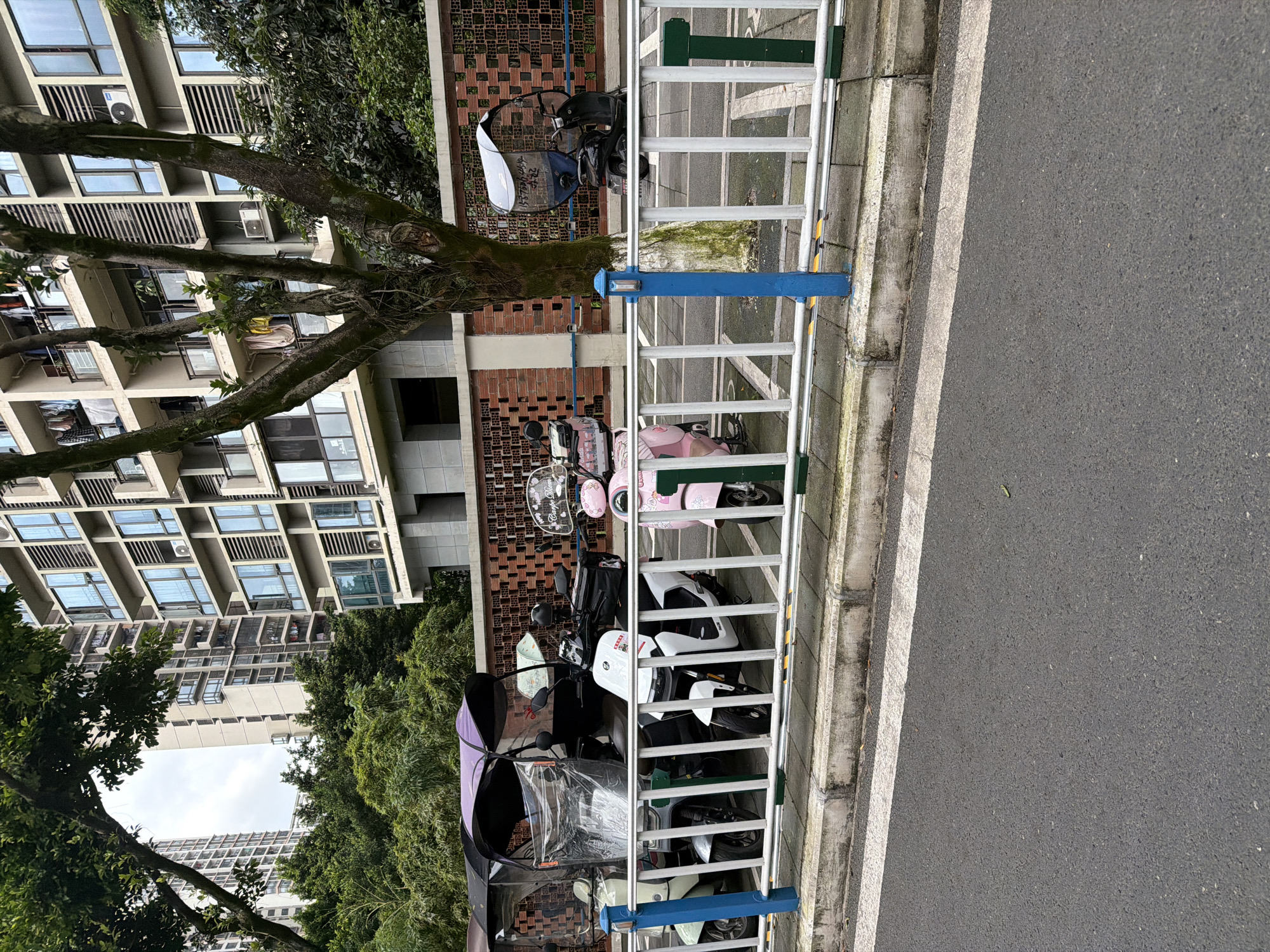 The image depicts a street scene in Chongqing High-tech Zone, China. A multi-story apartment building is visible in the background, partially obscured by trees. In the foreground, several electric motorbikes are parked behind a white and green metal fence with a blue post. The road is asphalt, with a white line marking the edge. The bikes are of various colors and models, some equipped with rain covers. The fence separates the parking area from the roadway.