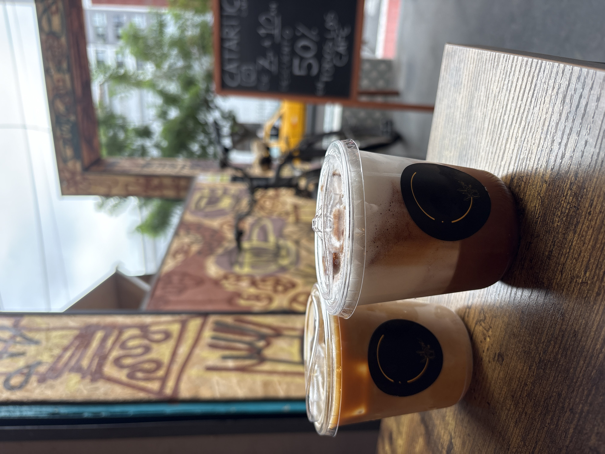 This is a medium-shot, eye-level photograph taken outdoors, likely in San Isidro, Peru, focusing on two iced coffee drinks on a wooden table. The drinks are in clear plastic cups with clear lids, each featuring a black circular logo with a golden outline and design. The cup on the left contains a light brown beverage, while the cup on the right has a darker brown bottom layer and a foamy, lighter brown top layer, suggesting it's a layered coffee drink.
The background is slightly blurred but reveals elements of a city street. To the left, there's a wall with a colorful, abstract mural. Behind the drinks, a black bicycle is visible, and further back, a yellow taxi is parked on the street. A wooden signpost with black lettering is positioned to the right, with "CATARTIC CAFE" prominently displayed at the top, along with pricing and a "50%" discount offer, indicating this is an outdoor seating area of a cafe. Trees and buildings are also visible in the distance. The lighting suggests an overcast day. No people are clearly visible in the scene.