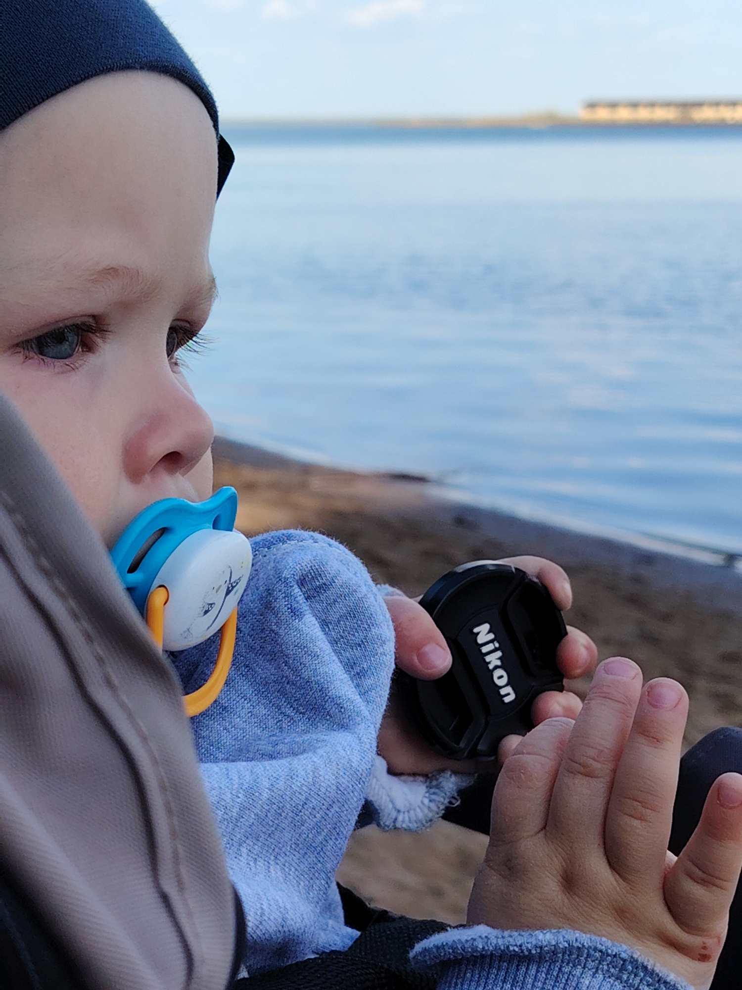 A child with a blue pacifier is seated and holding a Nikon lens cap. The scene appears to be on a beach, with a body of water and a structure visible in the background. Based on the presence of a structure resembling the Nizhnekamsk hydroelectric power plant, it is highly probable that the location is in the city of Chaikovsky, Russia.