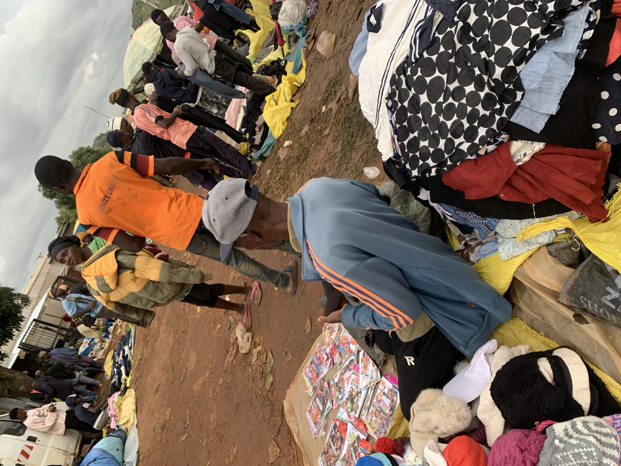The image depicts an outdoor market scene in Gura Top, Nigeria, with an overcast sky. In the foreground, a man wearing a blue jacket with orange stripes and a grey cap sits on the ground, looking at his phone. To his left are various items, including stacks of magazines, hats, and folded clothing. Behind him, a woman in a yellow and green jacket walks away from the camera, while other people are seen browsing or interacting in the market. The ground is predominantly dirt, with some areas covered by tarps and piles of clothing. The market appears to be selling a variety of goods, including clothing and possibly entertainment items like magazines.