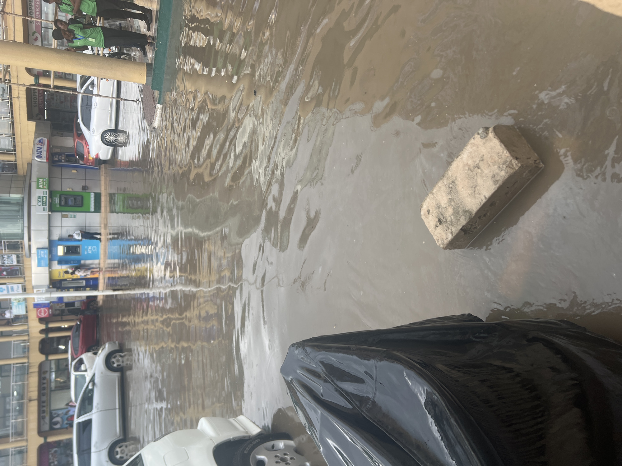 The image captures a scene of urban flooding in Dar es-Salaam, Tanzania, likely following heavy rainfall. The ground is largely submerged under murky brown water, reflecting the sky and nearby buildings.
In the immediate foreground, a large, black, reflective object, possibly a tarp or plastic covering, occupies the bottom right corner. To its left and slightly further into the scene, a light-colored brick or concrete block is partially submerged in the floodwater. The water shows ripples and movement, indicating depth and reflections.
Along the left side of the image, a commercial street or shopping area is visible. Several buildings line the thoroughfare, featuring storefronts with visible signage, including "SHOPPERS SUPERMARKET" and "ATM" machines belonging to "CRB BANK" (partially visible). Parked and possibly moving vehicles, predominantly white cars and one red car, are seen with their tires and lower parts of their bodies submerged in the floodwaters.
On a raised, dry area next to the buildings on the upper left, several people are standing. A few individuals in green shirts and dark trousers are clearly visible, appearing to observe the flooded street. The scene suggests a disruption to daily life caused by the inundation, though people and vehicles are still present in the area. The time of day appears to be daytime, with bright light creating strong reflections on the water's surface.