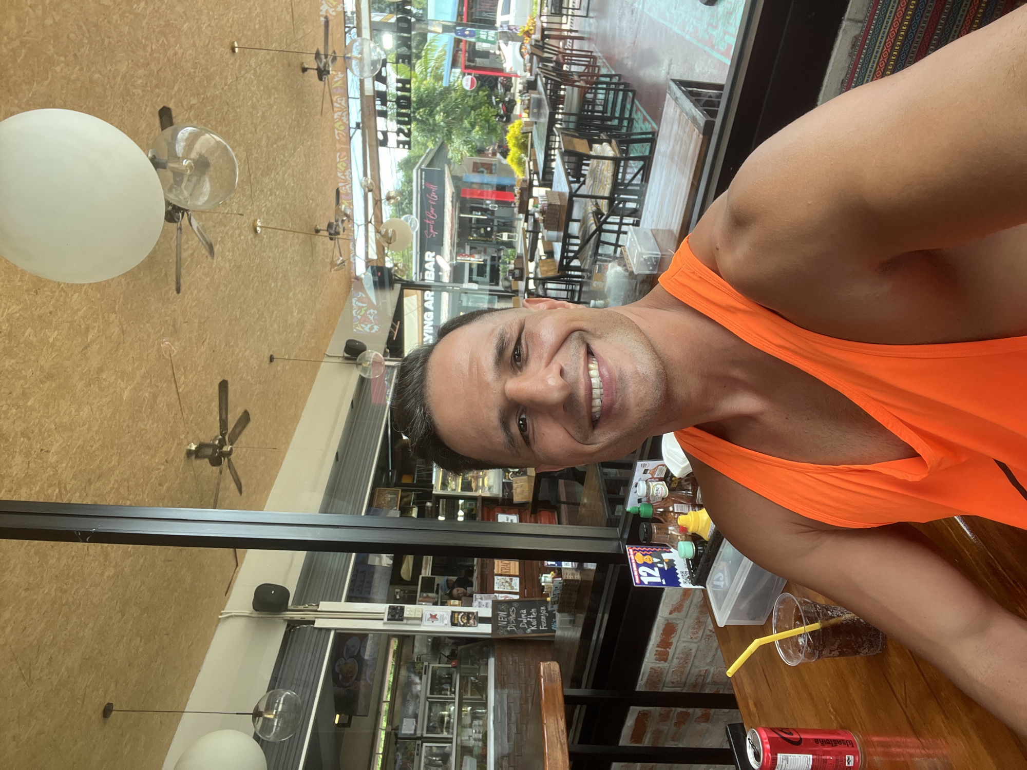 A man with short, dark hair and a tan complexion is taking a selfie. He is wearing an orange tank top and smiling at the camera. He is seated at a wooden table in an outdoor restaurant or bar. In front of him on the table is a plastic cup with a dark beverage and a yellow straw, a red can of Coca-Cola, and a clear plastic container. Behind him, through a large window, a bustling street scene in Chalong, Thailand is visible. Several tables with chairs are set up in the restaurant, and ceiling fans are visible on the ceiling. The overall atmosphere appears casual and lively.