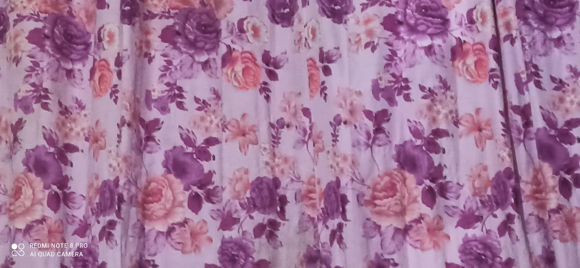 A floral patterned fabric, possibly a curtain or drapery, is displayed. The pattern features a dense arrangement of various flowers, predominantly in shades of purple and pink, with some lighter peach-colored blossoms. The fabric exhibits vertical folds, suggesting it is hanging. The bottom left corner of the image contains a watermark indicating "REDMI NOTE 8 PRO AI QUAD CAMERA." The specific location in Sedati Agung, Indonesia, is not visually determinable from the provided image and crops; the image itself is a close-up of a textile pattern.