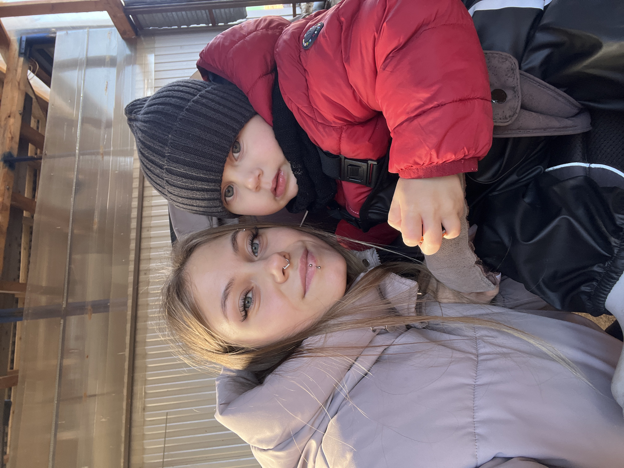 The image shows a young woman and a toddler posing for a selfie outdoors. The woman is lying on her back, wearing a light purple puffer jacket and light-colored hair. The toddler is on top of her, wearing a dark gray beanie and a red puffer jacket. The location appears to be an outdoor structure in Фоки, Russia, as suggested by the prompt, based on the visible construction and siding.