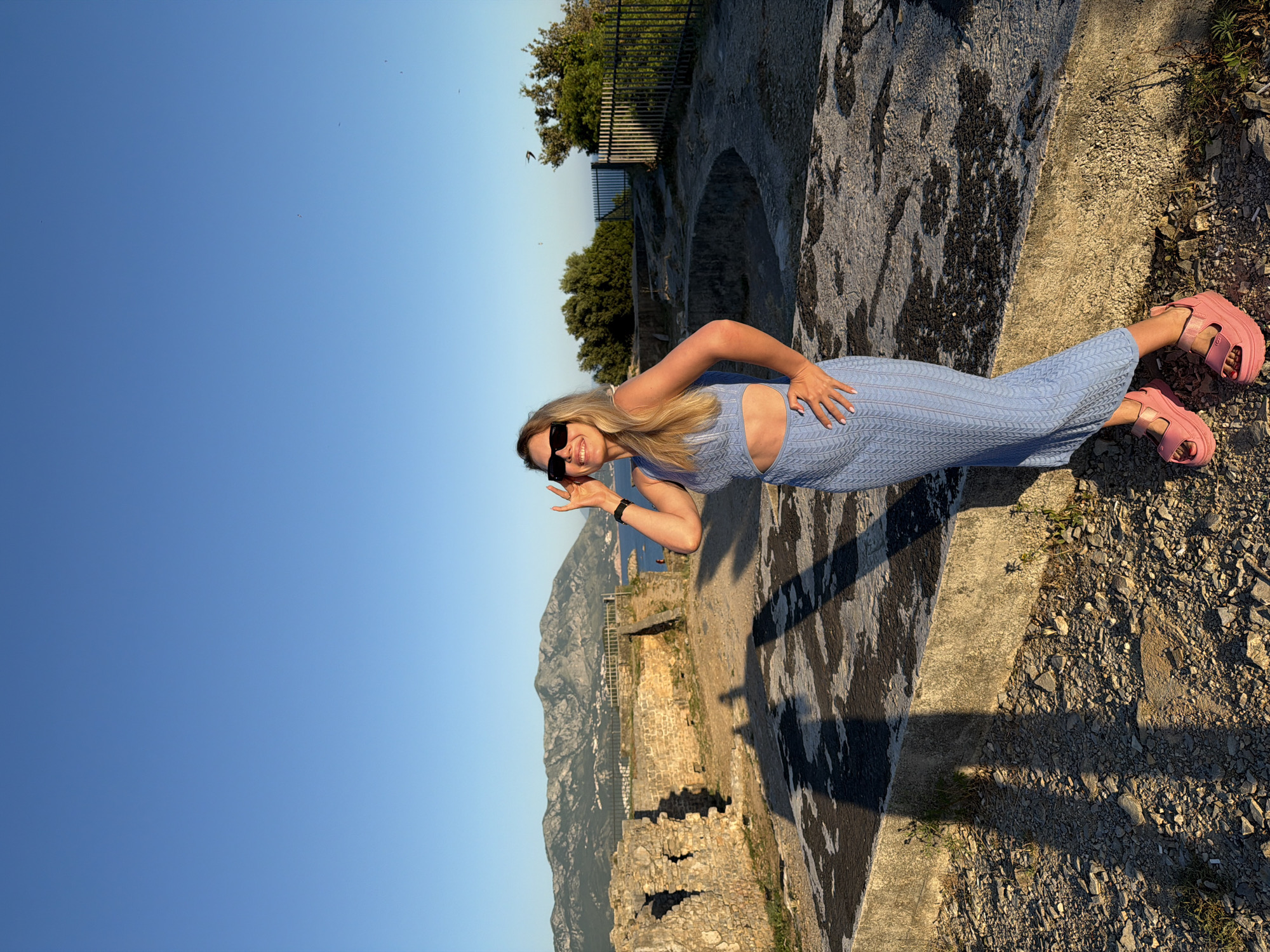 A fair-skinned woman with long blonde hair, wearing black sunglasses, a light blue knitted two-piece outfit (a crop top and a matching midi skirt with a side cutout), and bright pink platform sandals, stands smiling at the camera. Her left hand is raised towards her sunglasses, and her right hand rests on her hip. A dark watch is visible on her left wrist.
The setting is an outdoor, elevated location featuring ancient stone ruins and a concrete platform. The ground is covered with gravel and dirt, with sparse vegetation. In the background, a vast body of blue water, likely the Adriatic Sea, stretches towards a distant mountainous coastline dotted with white buildings, consistent with the landscape of Budva, Montenegro. To the right, green trees and a dark metal fence are visible. The sky is clear and bright blue. Long shadows are cast on the ground from the left, indicating the sun is low, suggesting either early morning or late afternoon light. Several small, dark specks, possibly birds, are visible in the upper right quadrant of the sky.