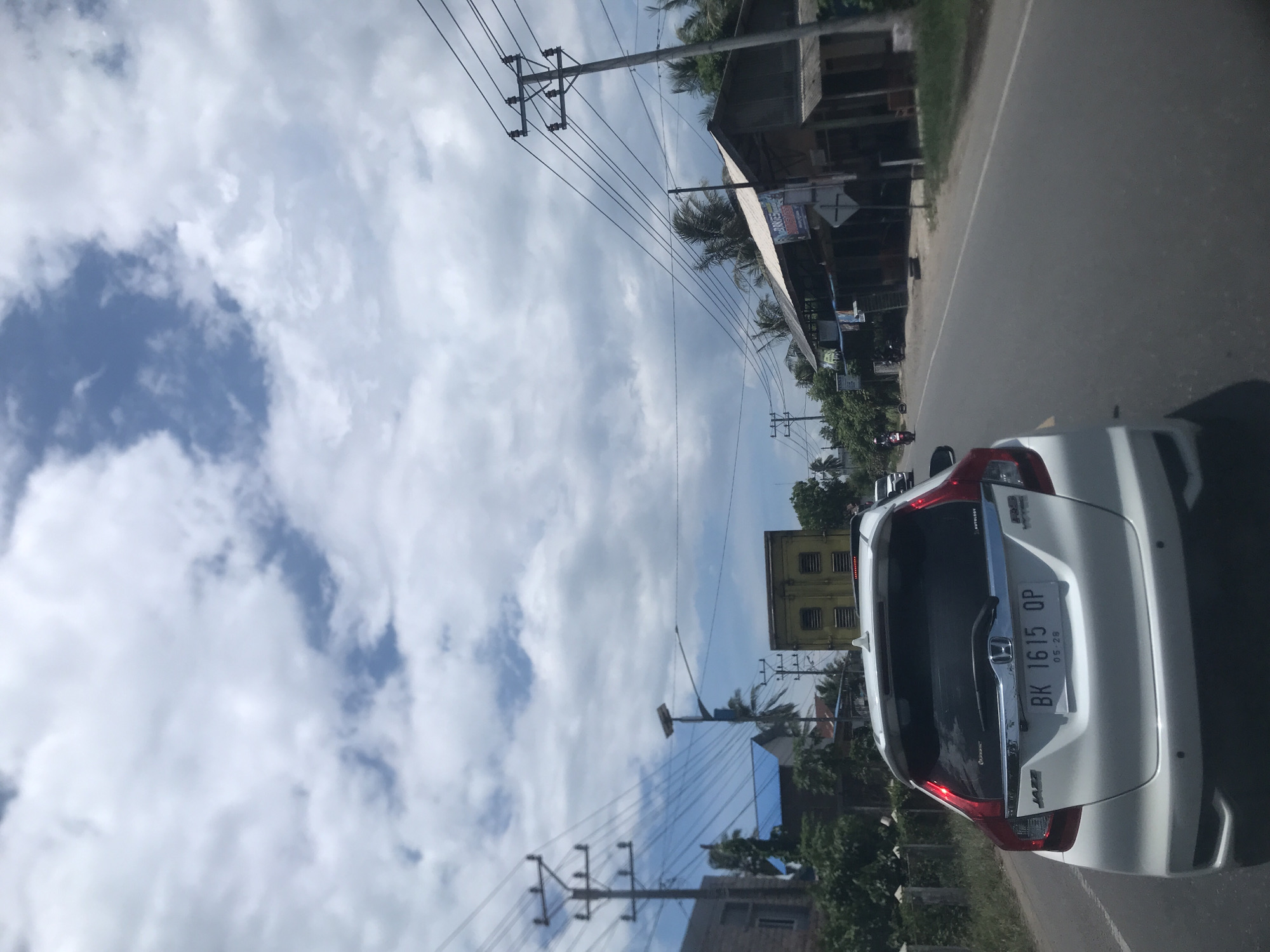 The image shows a street scene in Lhokseumawe, Indonesia, taken from inside a vehicle looking to the right. A white Honda Jazz car is partially visible in the foreground, its license plate reading "BK 1615 OP." The car appears to be moving along a road. On the side of the road, there are utility poles with power lines running above. Buildings and vegetation line the road. The sky is visible above, featuring clouds. The weather appears to be sunny.