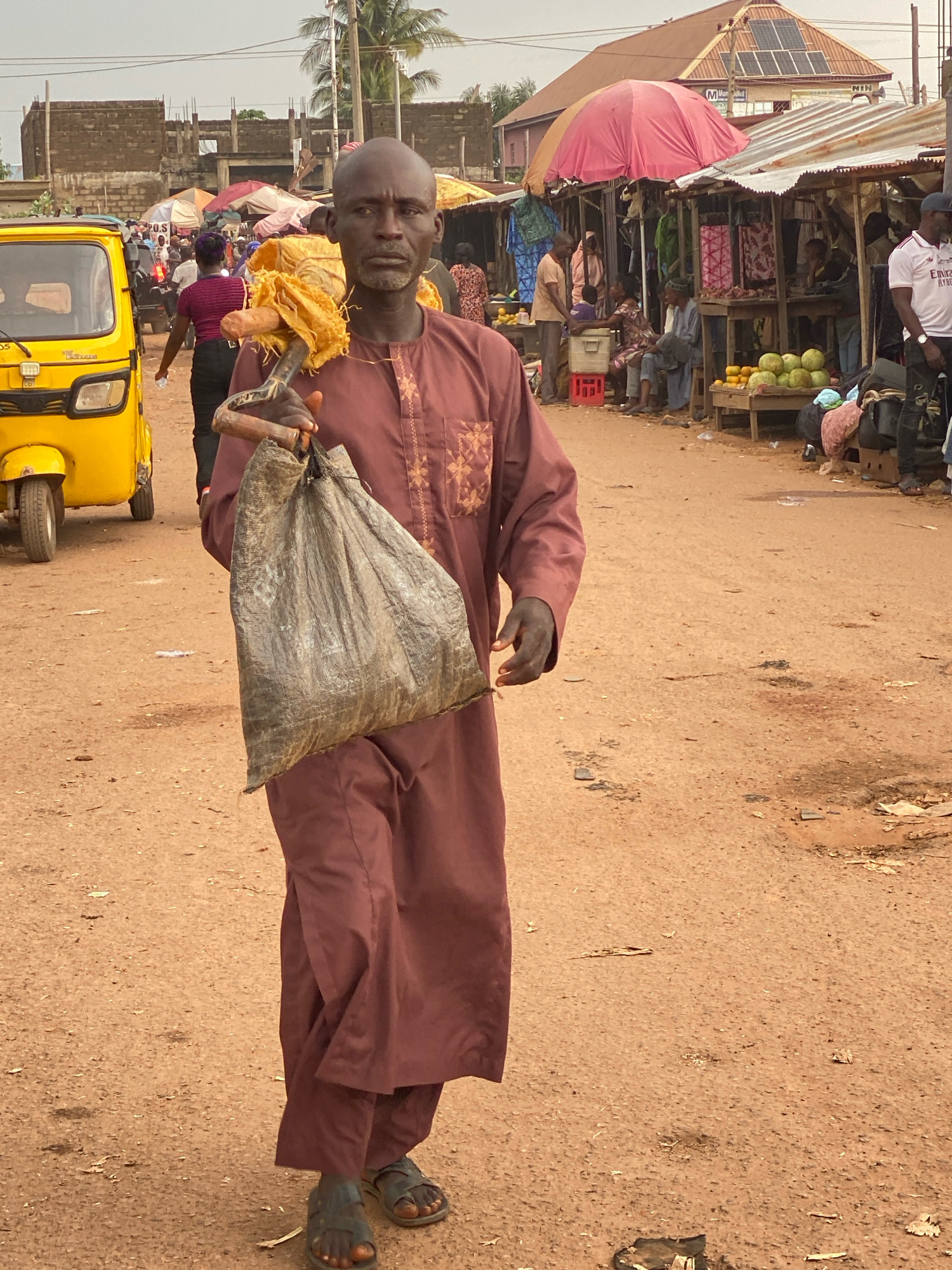 A man in a maroon traditional robe walks through a busy market in Sabon Tasha, Nigeria, carrying a large, dirty sack over his shoulder. He is holding a stick, likely used to help balance the sack. His face is visible, showing a neutral expression. In the background, a yellow auto-rickshaw is parked, and various market stalls with awnings display goods such as fruits and textiles. People are milling about, engaged in market activities. The ground is dusty and uneven, typical of a busy outdoor market. The sky is overcast, suggesting a cloudy day.
