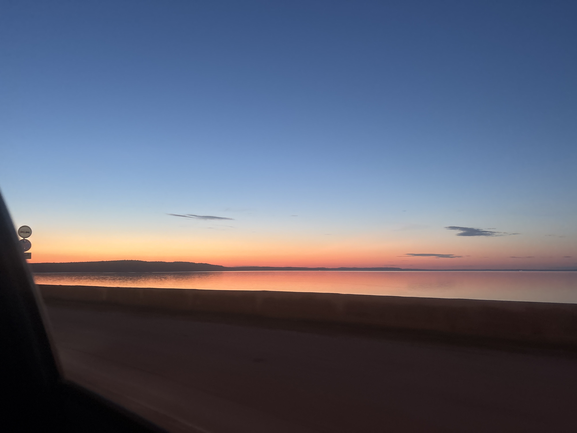 This image presents a serene twilight landscape in the city of Chaikovsky, Russia, likely captured from a moving or stationary vehicle. The upper two-thirds of the frame are dominated by a clear sky, transitioning from a deep cerulean blue at the top to vibrant oranges, reds, and pinks at the horizon, indicating either sunset or sunrise. Several small, wispy clouds are present in the blue sky and near the colorful horizon.
Below the horizon, a calm body of water, likely a large lake or river, extends across the mid-ground. The water's surface reflects the sky's vivid hues, creating a prominent band of orange and pink light stretching towards the viewer. A dark, low-lying landmass, consistent with a distant shoreline or hills, is silhouetted against the colorful sky on the far side of the water.
The foreground consists of a dark, flat surface, resembling a road or paved area, occupying the bottom quarter of the image. On the far left, a dark, undefined object, possibly a car's interior pillar or dashboard, partially obstructs two vertically stacked, circular road signs. The signs are mostly white with dark symbols, but insufficient detail is available for specific identification. No individuals, animals, or explicit actions are discernible within the frame. The scene is characterized by natural lighting and an expansive, tranquil environment.