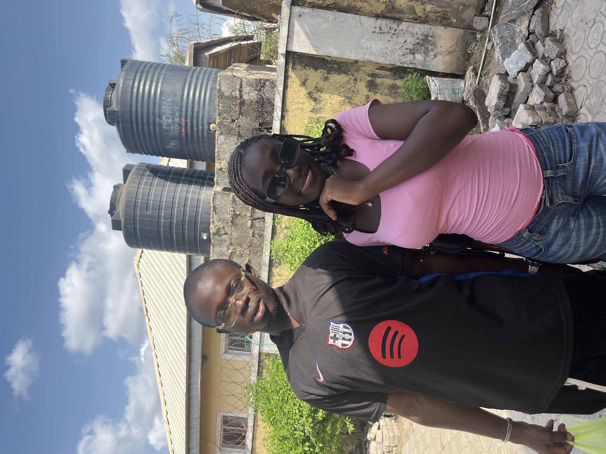 Two individuals, a male and female, are positioned outdoors with various structures visible in the background. The woman is wearing sunglasses, a pink shirt, and jeans. The man is wearing glasses and a black FC Barcelona shirt with a Spotify logo. There are two large, black water tanks situated near a concrete structure. A yellow building with barbed wire is also visible. The image was taken in Abuja, Nigeria.