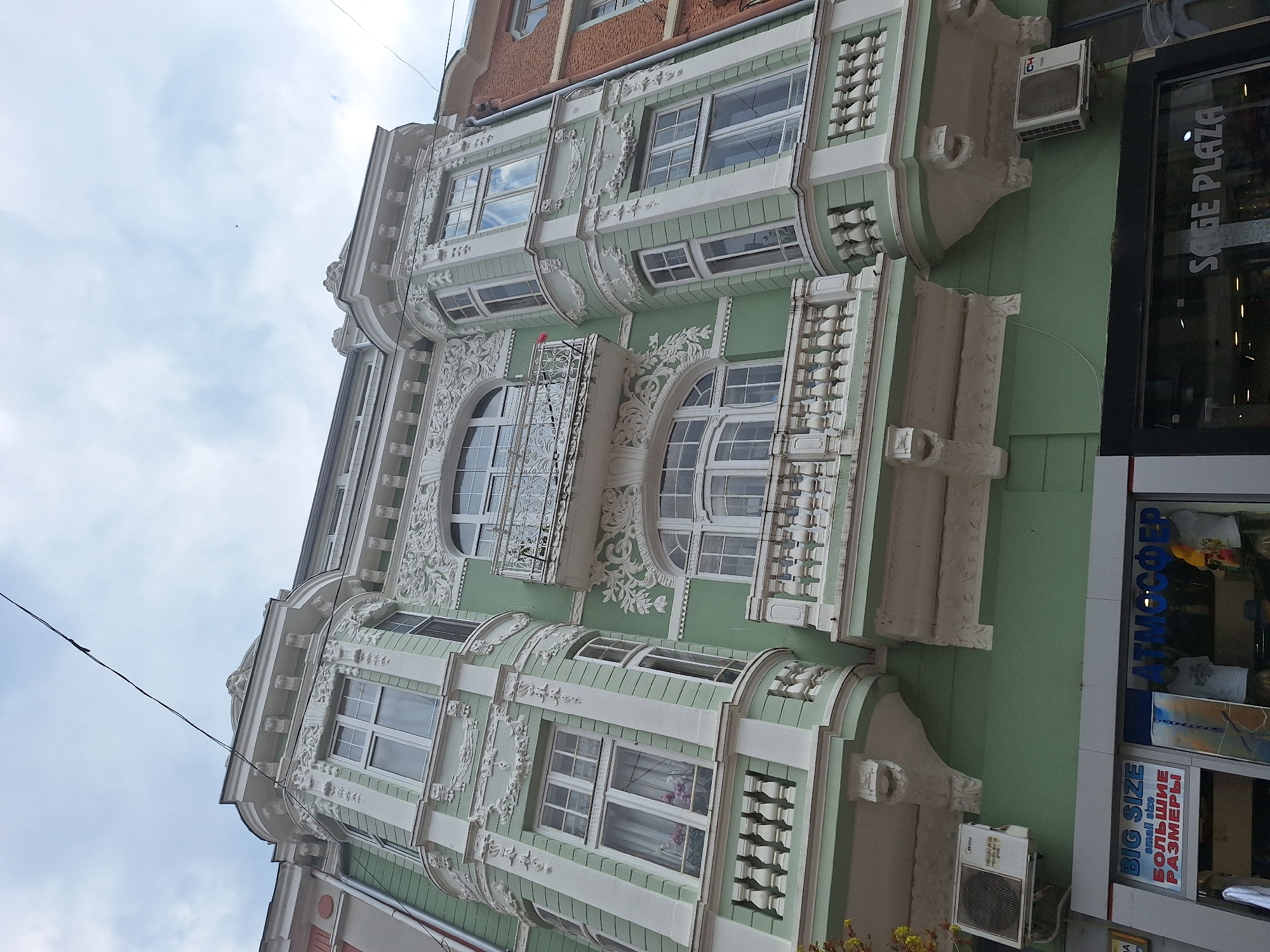 The image depicts a building facade in Varna, Bulgaria. The building features neo-baroque architecture with ornate white detailing on a green background. The facade includes arched windows, some with small balconies and decorative ironwork. There are also rectangular windows, some of which have air conditioning units mounted below. A storefront with signage, including the words "Sage Plaza" and Cyrillic text promoting "Big Size" clothing, is partially visible on the right side of the image. Overhead electrical wires are present in the top left corner, visible against a cloudy sky.