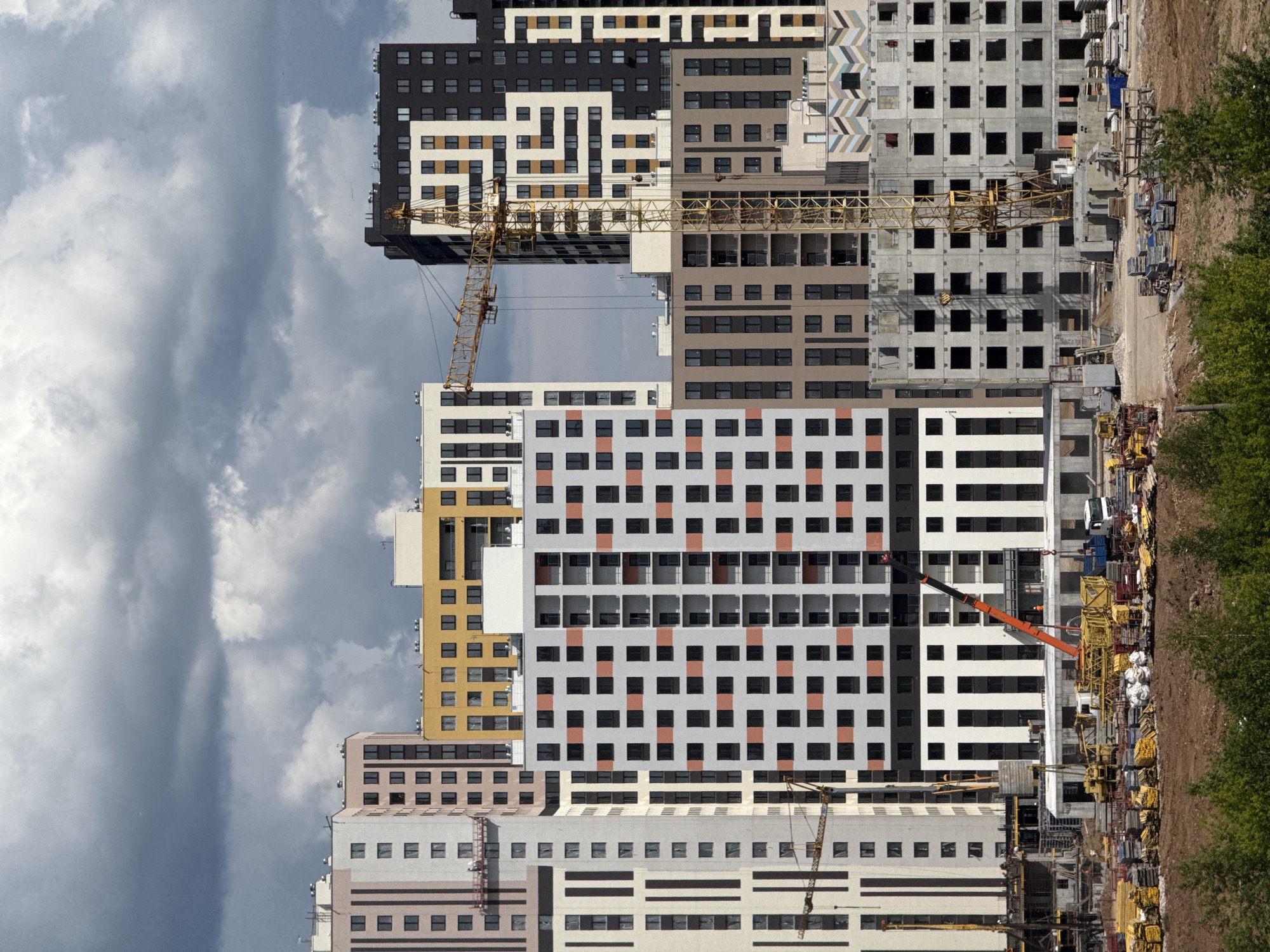 A large-scale construction site is depicted under an overcast sky, filled with multiple tall, modern residential buildings in various stages of completion. The sky is largely covered with dense, light-grey clouds.
Several multi-story apartment buildings dominate the scene. Some have finished, patterned facades in colors such as black, white, beige, grey, and orange, featuring grids of windows and occasional balconies. Other buildings are still in their skeletal concrete phase, revealing empty window openings and structural components.
Multiple yellow tower cranes are prominently positioned within and above the buildings, signifying active construction. One large crane arm stretches horizontally across the mid-section of the image, spanning different building structures. An orange and black mobile crane is also visible on the ground level to the right.
The right side of the image features the ground level of the construction site, with disturbed brown earth, dirt roads, and scattered construction materials like lumber, scaffolding, and bagged supplies. Green foliage, possibly trees or bushes, lines the far right edge of the construction area, suggesting an undeveloped or natural boundary.
The scene conveys a sense of a significant urban or suburban development project. No people are visible, and there is no discernible text on any objects or buildings. The time of day appears to be daytime, with natural, diffused light from the cloudy sky.