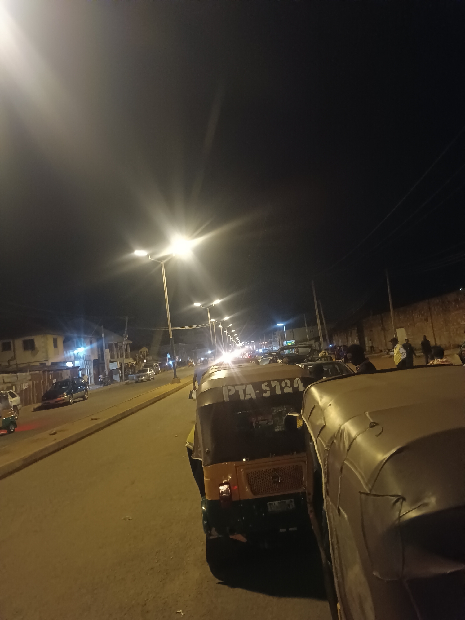 The image depicts a street scene at night in Bukuru, Nigeria. Streetlights illuminate the road, which is lined with vehicles, including "keke napeps" (auto rickshaws). People are standing and walking along the sidewalk and street. Buildings are visible in the background. One "keke napep" has license plate "PTA-5724".