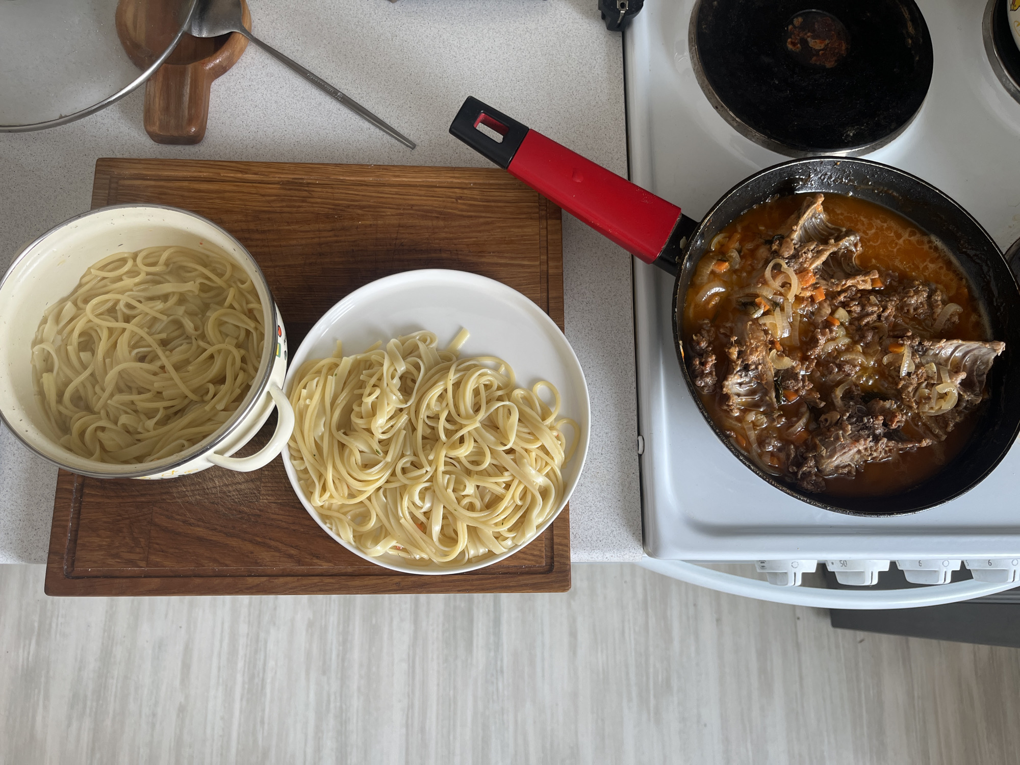 This is an overhead, close-up shot of a cooking scene. The image shows a stovetop with a white enamel finish and black electric burners. In a black frying pan with a red handle, pieces of meat are simmering in a reddish-brown sauce with visible onions and carrots. To the left of the pan, a white plate is piled high with cooked spaghetti. Behind the plate, a wooden cutting board holds a pot also filled with cooked spaghetti. A metal spoon and a lid rest on a wooden holder to the right of the cutting board. The overall impression is of a home kitchen setting, possibly in Noyabrsk, Russia, with ingredients prepared for a meal.