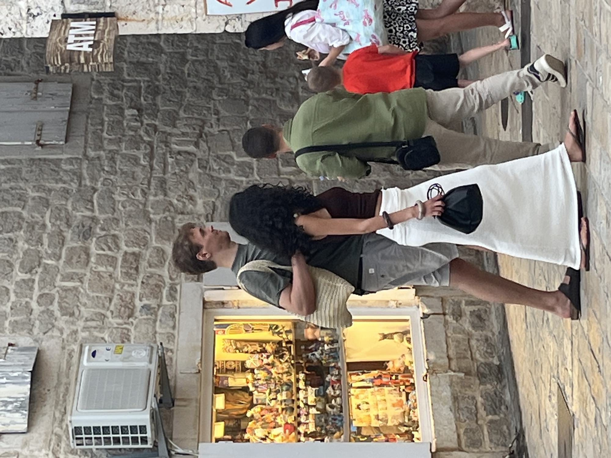 The image shows a street scene in Budva, Montenegro. A group of people are walking on a cobblestone street in front of a building with a stone facade. On the left, a shop window is filled with various figurines. An air conditioning unit is mounted on the exterior wall above the shop. Further to the right, a wooden sign with "ATM" is visible.
The individuals in the foreground include a man in a green t-shirt and grey shorts, carrying a bag over his shoulder. Beside him, a woman with dark curly hair wears a brown tank top and a long white skirt, holding a black clutch. Behind them, a man in a green shirt and light-colored pants walks with a black bag slung across his chest. A young boy in a red t-shirt and black shorts is also present, and a woman in a white shirt and a patterned skirt is visible further back, carrying a tote bag. The people appear to be walking away from the camera. The overall atmosphere suggests a warm day, with the people dressed in casual summer attire.