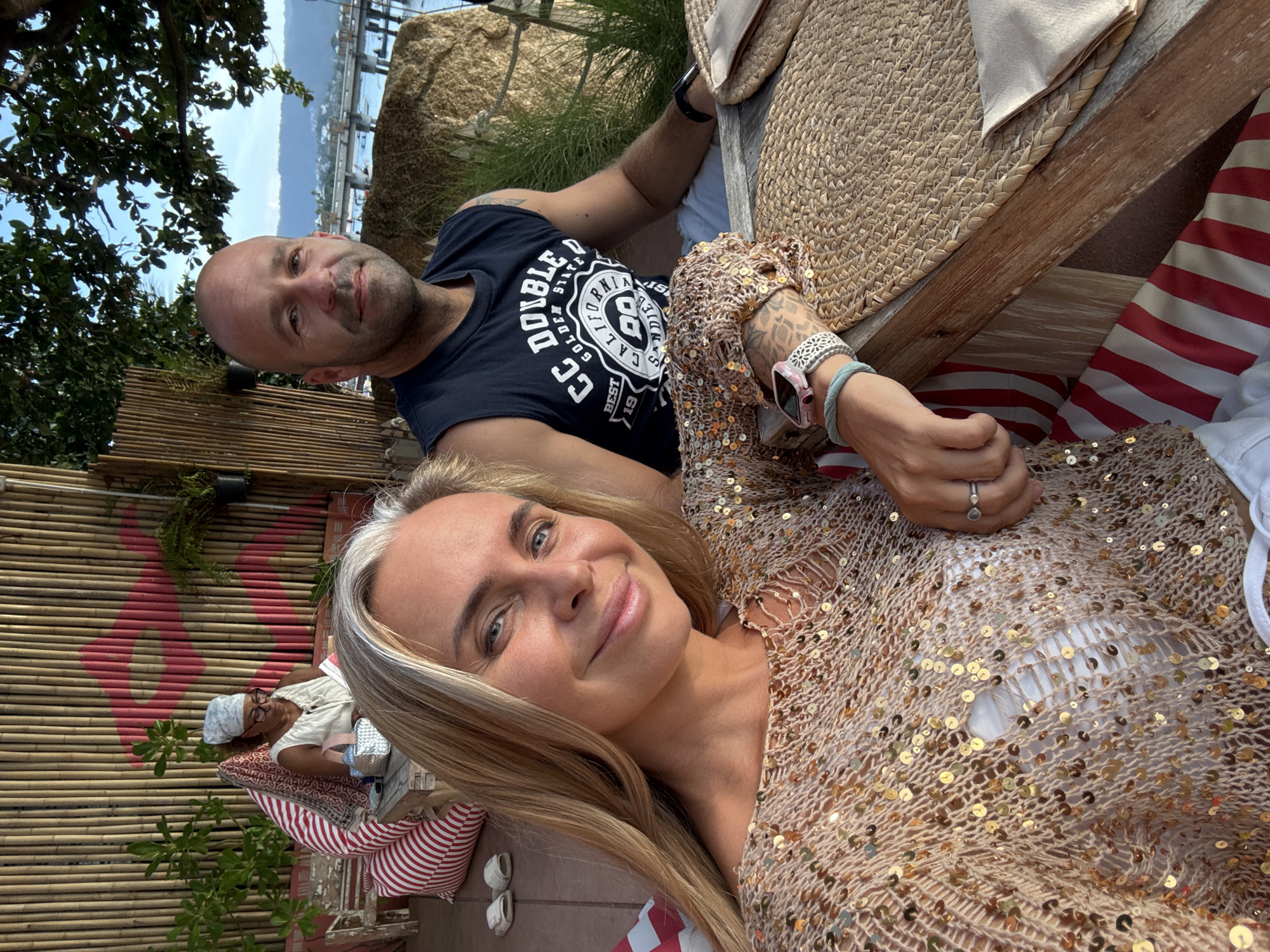 In Ko Samui District, Thailand, a man and a woman pose for a selfie in a relaxed outdoor setting. The woman, in the foreground, has blonde hair and is wearing a gold sequined top. Her arm, adorned with a ring and a watch, rests on the table. The man, slightly behind her, is bald and wears a dark sleeveless shirt. Behind them, a woman with glasses and a headscarf reads a book while seated on a striped cushion. The background features a bamboo wall, lush greenery, and a glimpse of the ocean with a pier. The table is set with woven placemats and napkins.