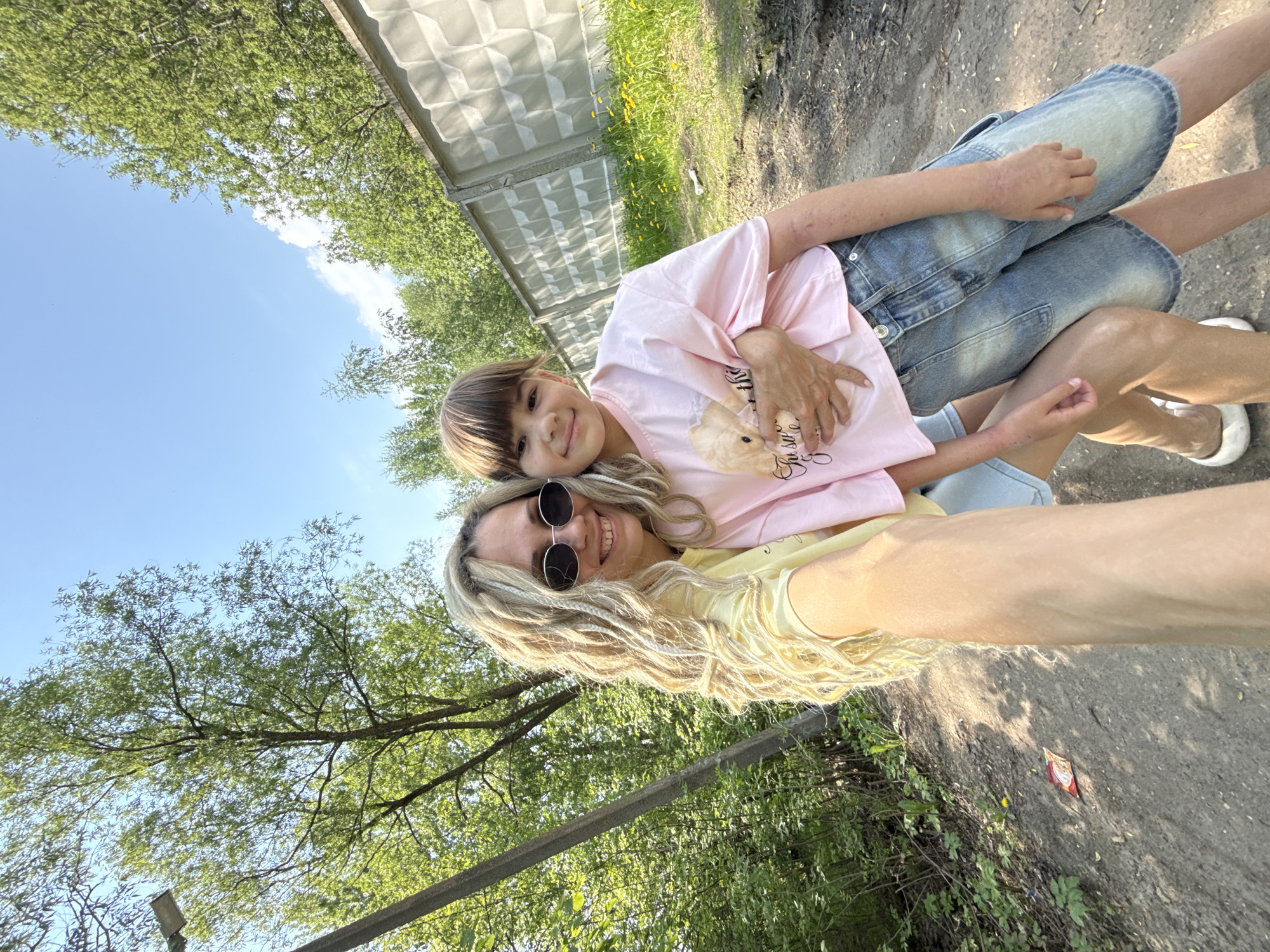 A woman with blonde dreadlocks and sunglasses is taking a selfie with a child. The child has brown hair and is wearing a pink shirt and blue shorts. They are outdoors, with trees and a blue sky visible in the background. A metal fence is also visible. The photo appears to be taken in Rusanovo, Russia, given the location provided.