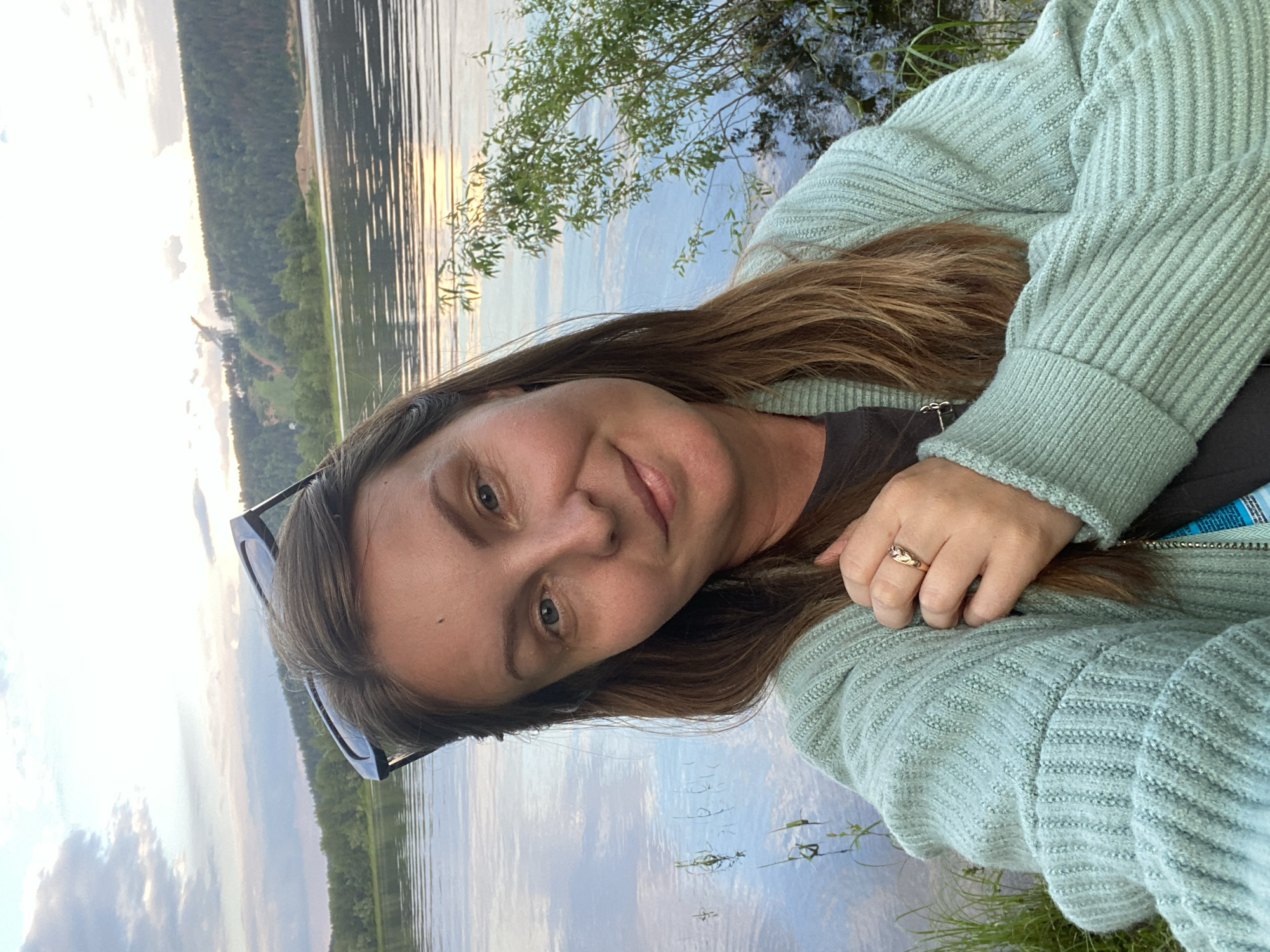 The media file is a portrait of a woman in an outdoor setting, likely near a body of water. The woman has long brown hair and is wearing a light green, ribbed cardigan over a dark shirt. She is looking towards the camera with a slight smile. A ring is visible on her right hand, which rests on her chest. She is also wearing dark-rimmed sunglasses perched on her head.
The background consists of a lake or river reflecting the sky, which is partly cloudy with hints of sunlight breaking through, suggesting it is either early morning or late afternoon. The far bank of the water is lined with trees and a green slope, with some buildings visible in the distance. Tall grass is growing in the foreground near the woman. The overall mood appears relaxed and serene, consistent with a peaceful outdoor excursion. There is no visible text in the image. The location context provided is Noviy, Russia.