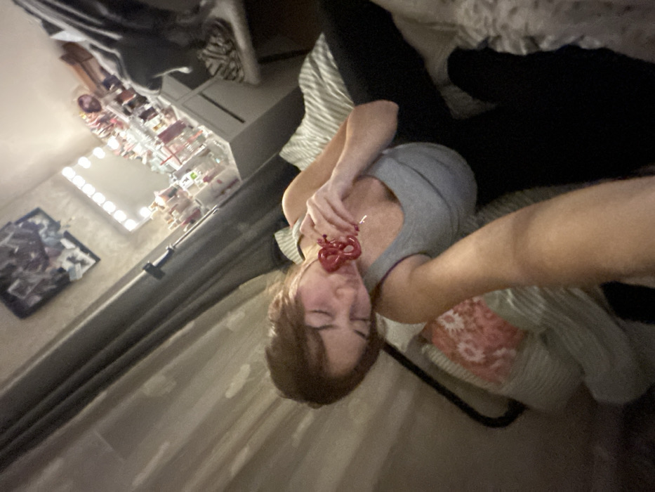 A woman lies on a striped bed or cushion, wearing a gray tank top and black leggings. She holds a red, heart-shaped object close to her face, her eyes closed. A vanity with lights and various cosmetic items sits against the wall to the left. The composition suggests she is taking a selfie with an extended arm. A framed image hangs on the wall nearby.