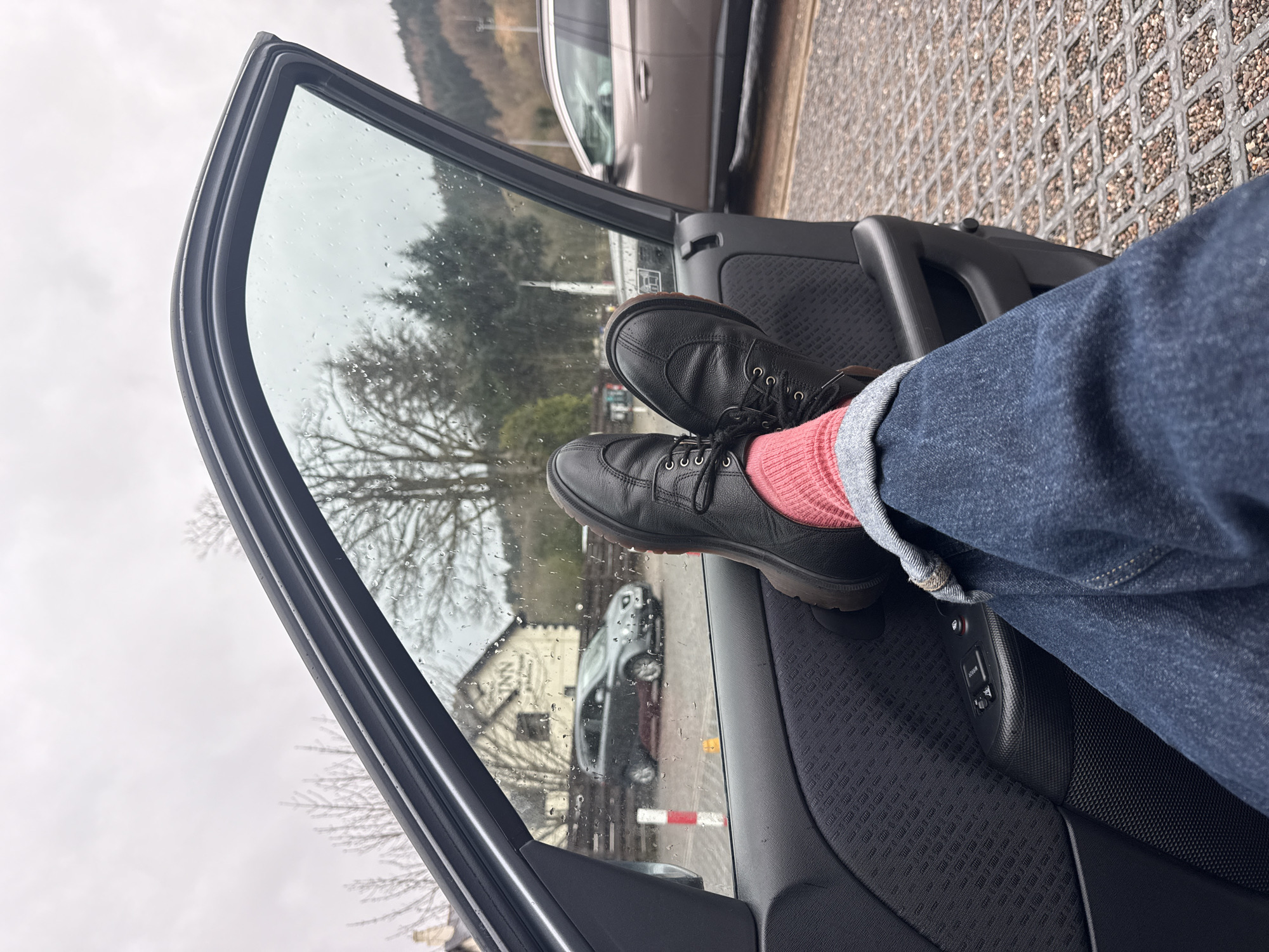 The image shows an interior view through the open door of a car, focused on a person's legs and feet resting on the door panel. The individual is wearing blue jeans cuffed at the ankle and pink socks paired with black leather shoes. Raindrops are visible on the car window. The background visible through the window includes a house and vehicle, and an overcast sky. The setting appears to be an urban or suburban area.