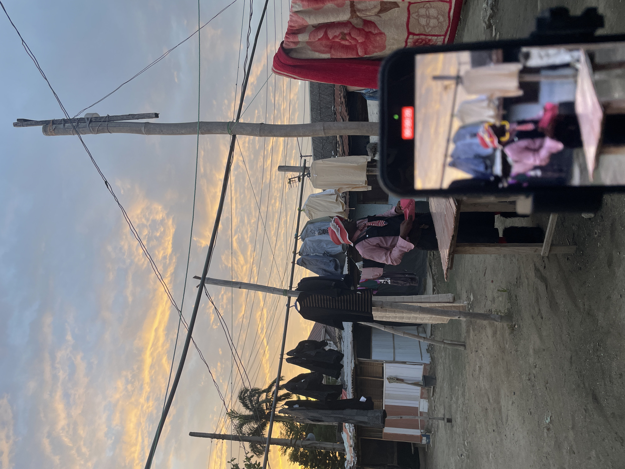 This media file, captured in Ado, Nigeria, depicts an outdoor scene at either sunrise or sunset, evidenced by the cloudy sky illuminated with orange and yellow hues. The image is oriented vertically (rotated 90 degrees clockwise from its natural horizontal view). In the foreground and midground, numerous clothes are hanging on lines to dry, strung between a series of tall, rustic wooden or bamboo poles. These poles are tied together with various ropes and wires, suggesting a makeshift setup. Overhead, several thin electrical wires crisscross the sky.
A notable element is a smartphone, positioned in the upper right, actively recording. Its screen clearly displays "REC" in red letters along with a red recording dot. The phone's display shows a blurred reflection or recording of clothes and parts of the surrounding environment. Above the clotheslines on the right, a floral-patterned blanket is draped. Beyond the clotheslines, simple structures, some appearing to be made of corrugated metal and wood, form the background, indicative of an informal settlement or rural area. A palm tree is visible on the left side of the frame, near the bottom. The ground beneath is uneven, dusty earth. The scene portrays a slice of daily life, focusing on practical activities like laundry against a striking natural backdrop.