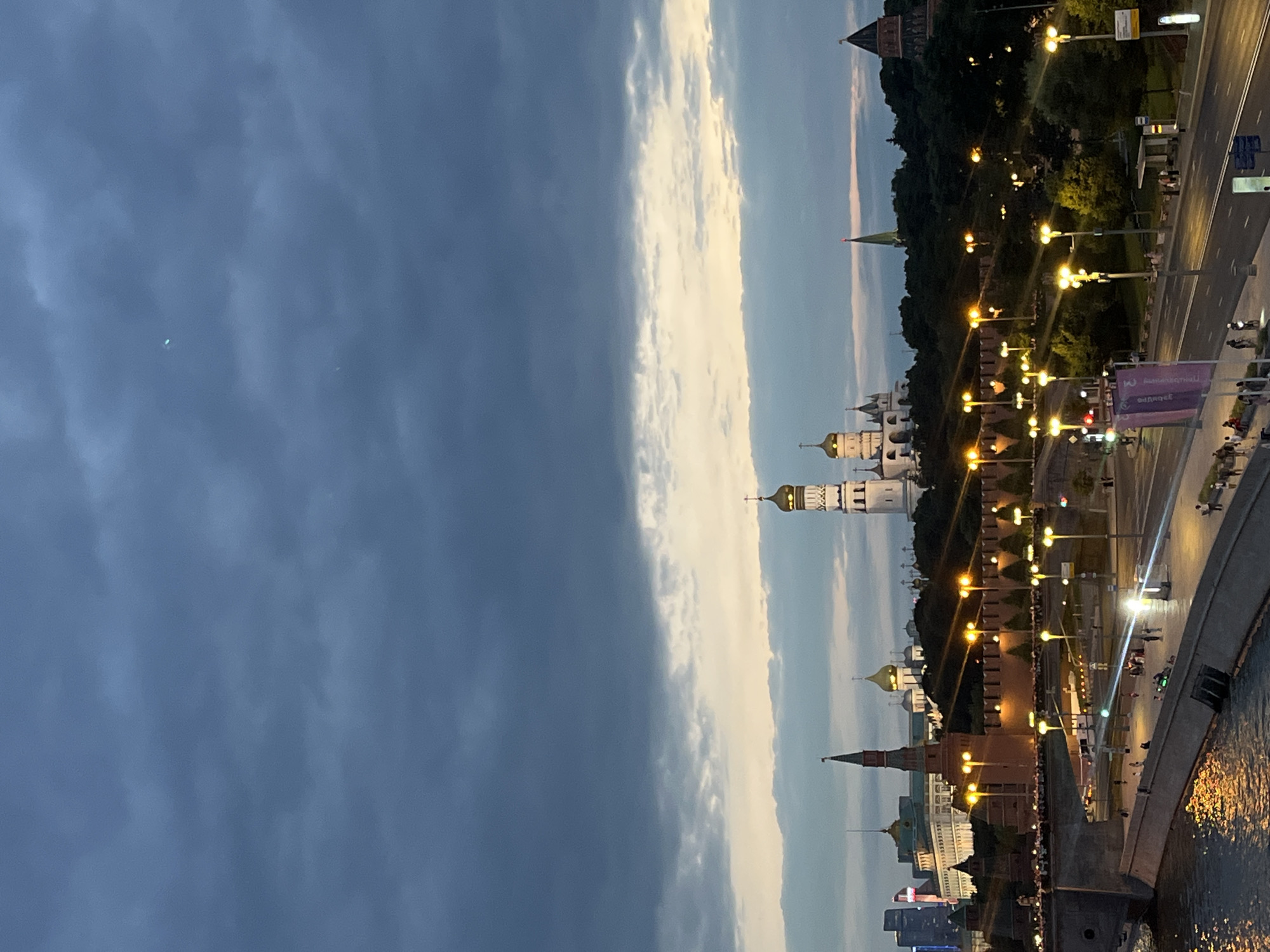 This is an exterior, eye-level, medium shot of the Moscow Kremlin at dusk. The sky is cloudy, with shades of blue and gray, and a hint of pastel colors where the sun is setting below the horizon. Several domes and spires of the Kremlin's cathedrals are visible, illuminated by artificial lights, as are the Kremlin walls and towers. In the foreground, a wide promenade is lined with streetlights, and a road with vehicles and pedestrians runs parallel to it. A body of water, likely the Moskva River, occupies the lower left corner of the image, reflecting the lights from the promenade. People are walking and standing on the promenade and sidewalk. The overall scene depicts a city scene at twilight, with historical landmarks and modern infrastructure coexisting.