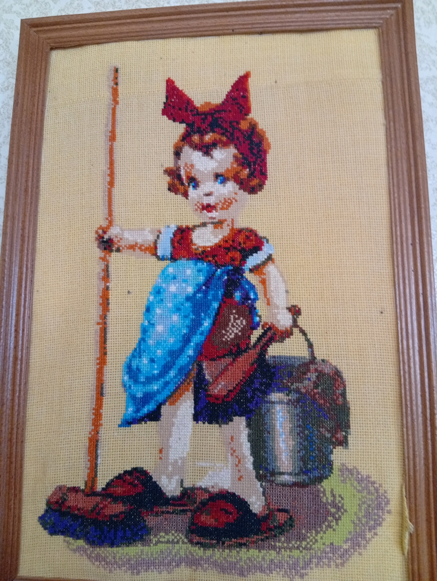 An unframed, framed, cross-stitch depiction of a young girl stands facing left, holding a broomstick in her left hand and a bucket in her right hand. The girl has blue eyes, rosy cheeks, and red lips, with auburn hair styled in pigtails tied with a red bow. She is wearing a red shirt with puff sleeves, a blue apron with white polka dots, and red shoes. A brown purse hangs from her right hip. The background is a pale yellow, with a patch of green and purple textured ground beneath the girl's feet. The overall scene suggests a child engaged in household chores. The image is presented on a neutral surface.