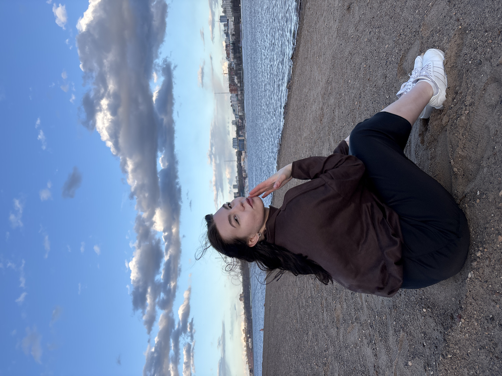 A young woman is depicted sitting or kneeling on a sandy and gravelly riverbank, posing for a photograph. She is positioned on the right side of the frame, facing slightly left and upwards with a contemplative expression, her left hand gently touching her chin. She has long, dark hair that appears slightly windblown. Her attire consists of a dark brown long-sleeved top, dark-colored leggings or trousers, and white athletic shoes.
To her left, a wide body of water, likely a river, stretches towards the horizon, displaying gentle ripples on its surface. Across the water, a cityscape with various buildings is visible along the distant bank, suggesting an urban setting such as Perm, Russia. The sky above is partly cloudy, with areas of bright blue sky interspersed with both wispy white clouds and larger, more textured grey clouds. The soft, natural light indicates either late afternoon or early evening. The ground she sits on is a mixture of fine sand and small pebbles. No visible text is present in the image.