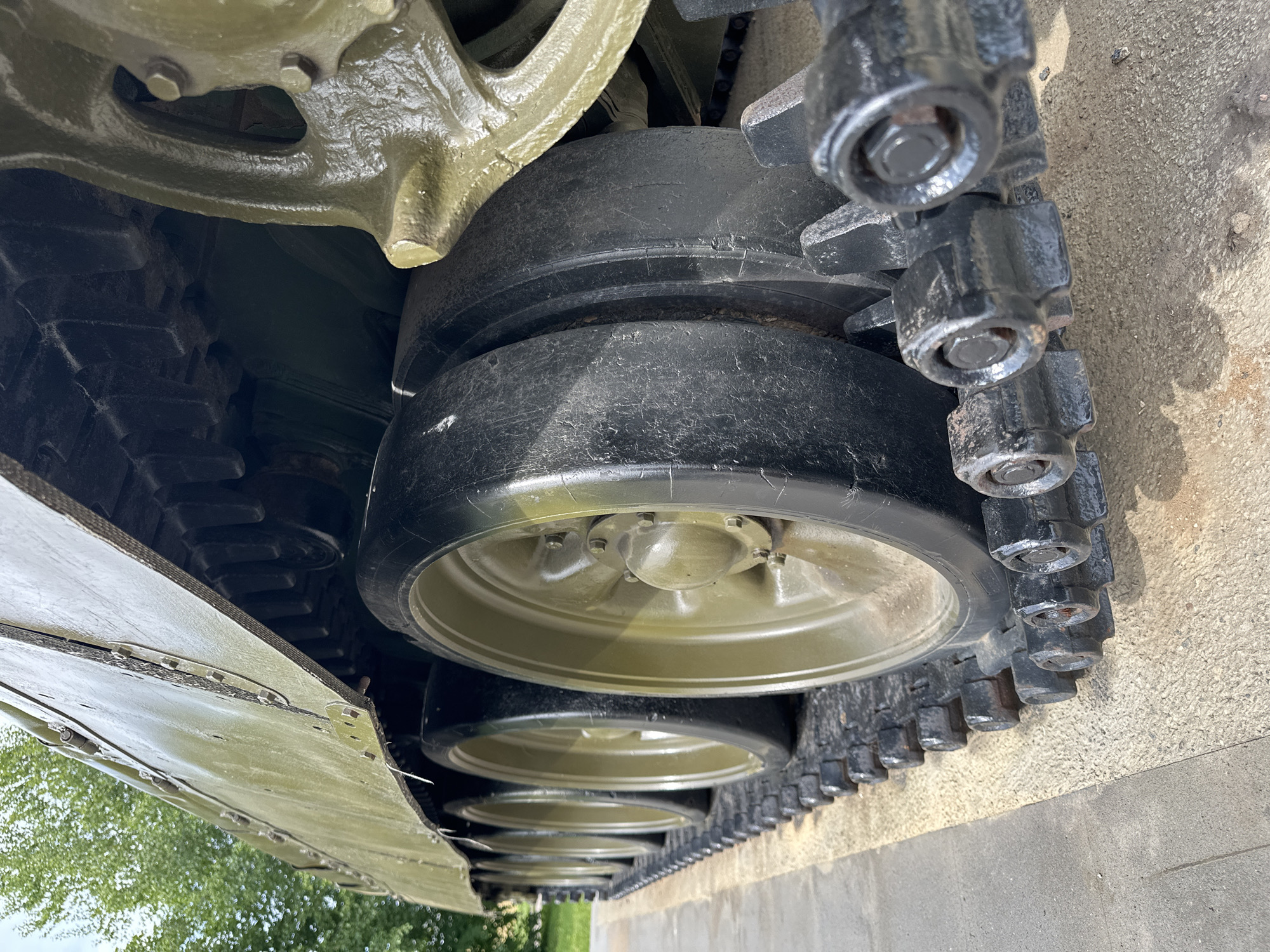 A low-angle, close-up shot focuses on the tracks and road wheels of a weathered olive-drab tank. The tank's metallic body, textured and marked with faint scratches, is visible at the top of the frame. Beneath it, a series of road wheels, painted a muted green, are partially obscured by thick, black tank treads. The treads, composed of interconnected metal segments, rest on a light-colored, granular surface, possibly a concrete or sand base. The lighting suggests an outdoor setting, with the tank positioned on a paved area adjacent to a patch of sand or gravel. The background is blurred, hinting at greenery and possibly trees, indicative of a park or outdoor exhibition space. The image is devoid of any human presence or activity.