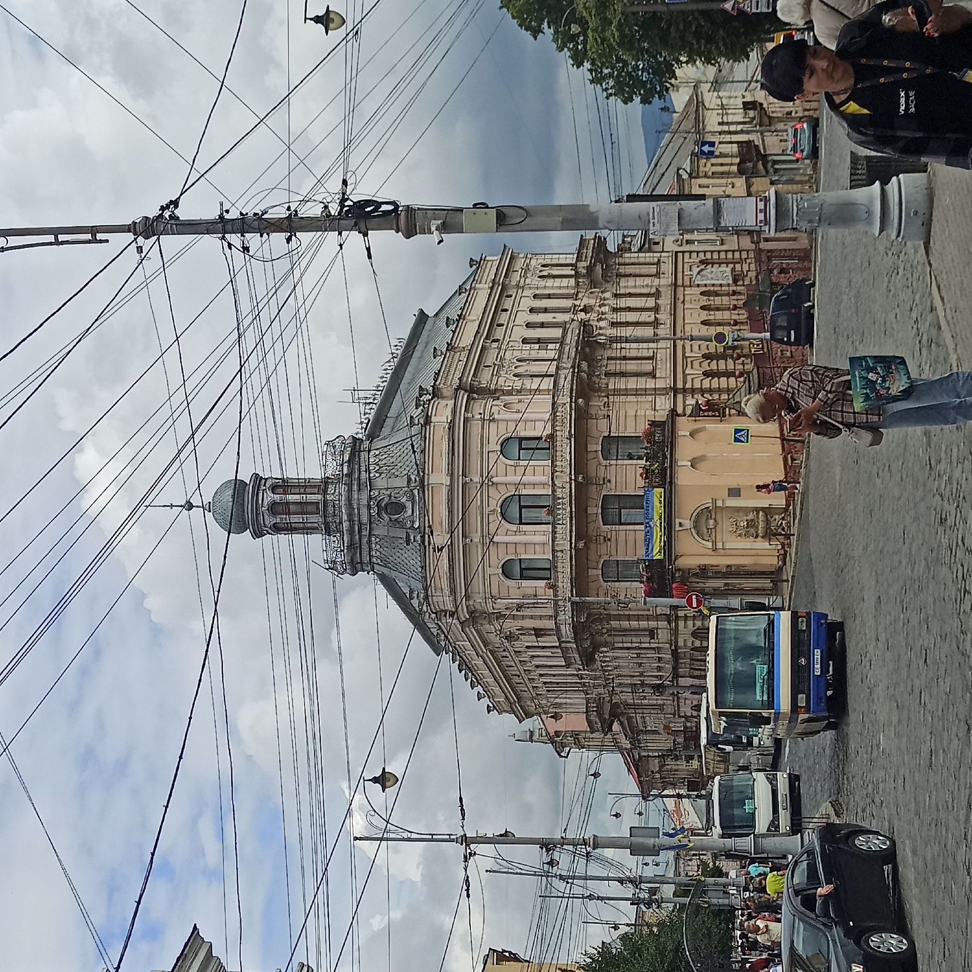 A street scene in Chernivtsi, Ukraine, shows a cloudy day with a mix of old and new architecture. A prominent, ornate building with a domed roof, likely a historic landmark, stands in the center, surrounded by other buildings with classical architectural elements. Overhead, a dense network of electrical wires crisscrosses the sky, supported by utilitarian poles. Several buses and a black car navigate the cobblestone street. Pedestrians are scattered throughout the scene, some walking, others standing. One woman in the foreground, holding a reusable shopping bag, is looking at her phone. Another person in a black jacket is walking past the viewer. A security camera is visible on a utility pole. A yellow and blue banner with Ukrainian text is displayed on one of the buildings. The overall impression is a bustling urban environment with a strong sense of history.