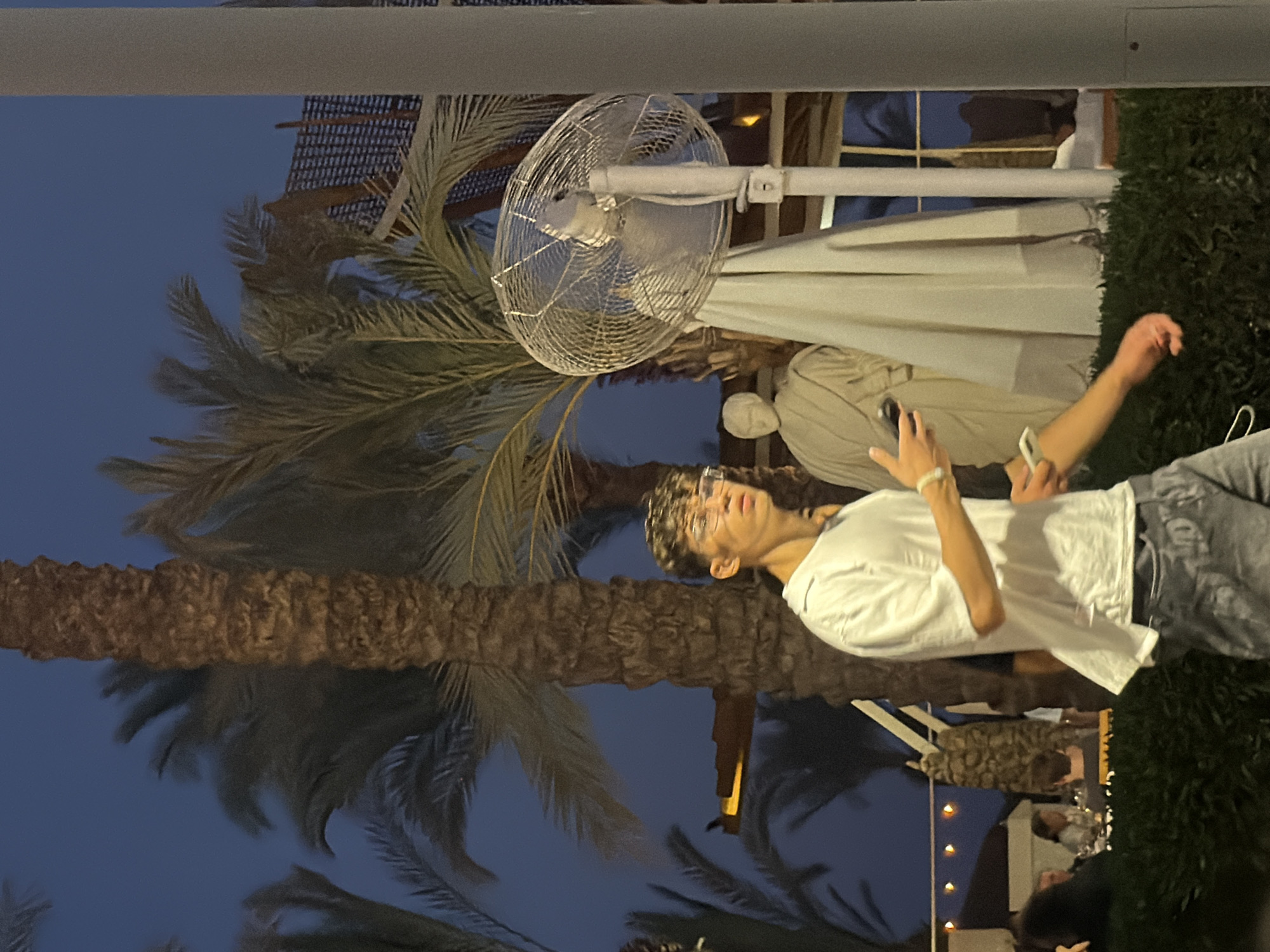 In Sharm El Sheikh, Egypt, a young man with curly hair and glasses, wearing a white t-shirt and grey pants, walks past palm trees. He holds a mobile phone in his left hand. In the background, there is a statue dressed in a toga and an industrial fan. Further in the distance, a restaurant setting is visible with people dining. The sky is a deep twilight blue.