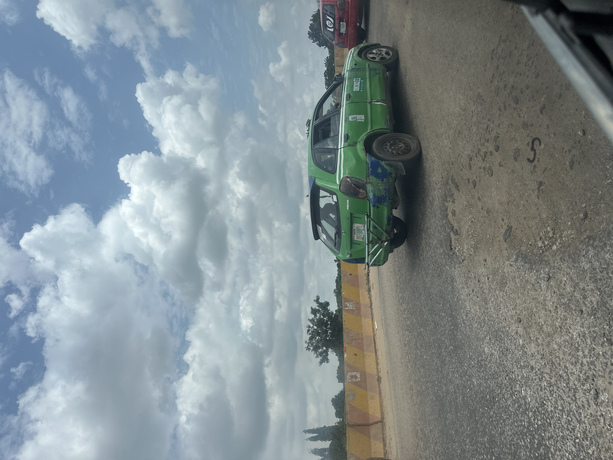 An exterior, eye-level, medium shot captures a green Mazda sedan with a white stripe on a paved road under a cloudy sky. The car, a taxi, has the license plate "LSD 109TL" and the number "000382" printed on its side. The vehicle appears weathered, with visible rust and damage on its rear bumper and lower body panels. In the background, a red car with a "PVL 5880" license plate is also visible, driving on the same road. The road itself is in disrepair, with visible potholes and cracks. A yellow and red barrier lines the left side of the road, separating it from a sandy embankment. Trees and foliage are visible in the distance, suggesting a semi-urban or rural setting. The clouds are large and white, scattered across a bright blue sky. The image was taken from inside another vehicle, as indicated by the blurry interior frame on the right side of the picture. The scene appears to be a typical day on a Nigerian road, likely within or near Abuja, given the license plate format.