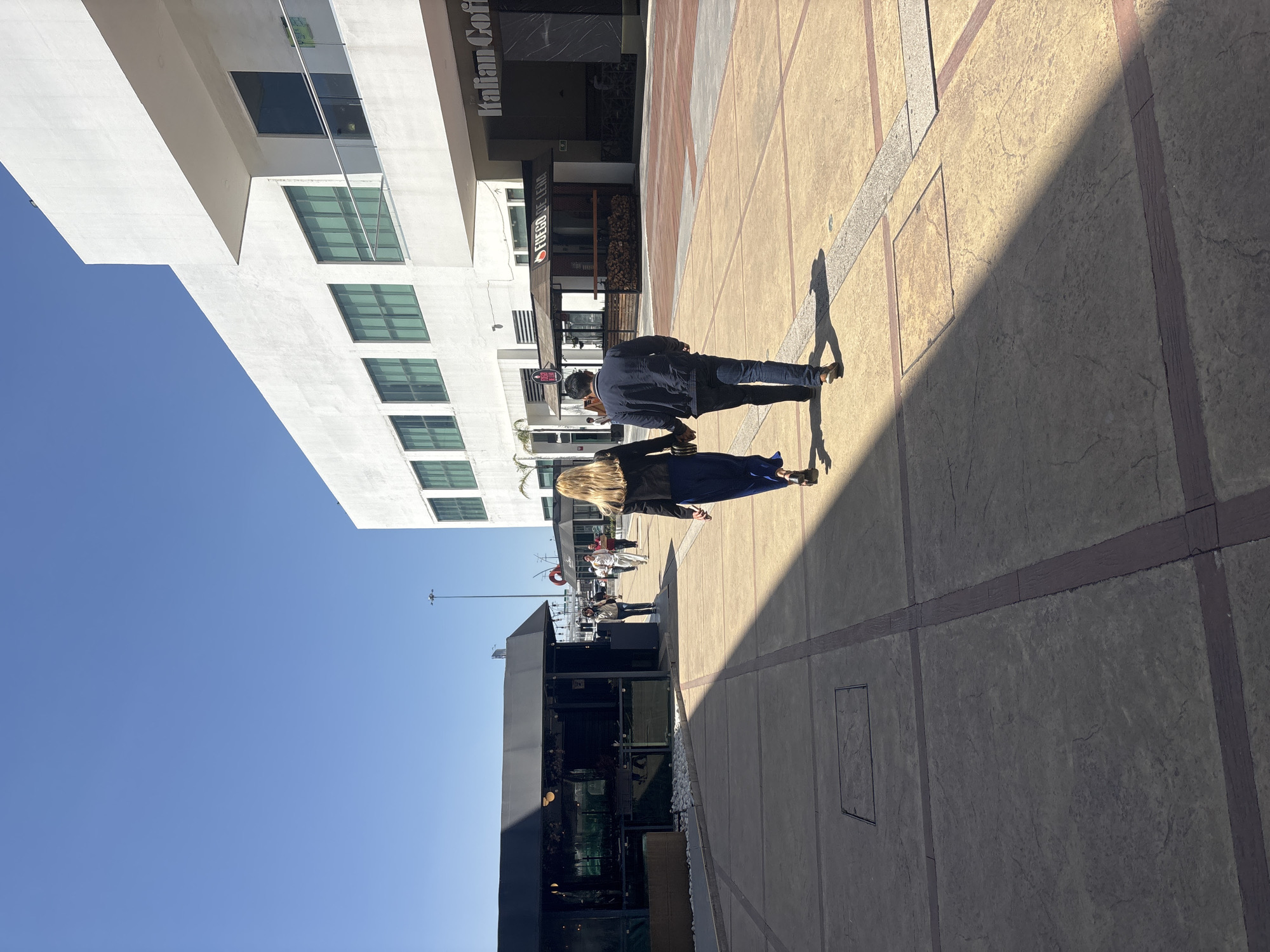 This image depicts an outdoor pedestrian walkway in a commercial area, likely part of or adjacent to the Angelópolis Lifestyle Center in Puebla, Mexico, under a clear, bright blue sky.
The scene is illuminated by strong sunlight, casting a prominent dark shadow diagonally across the light brown and dark brown grid-patterned pavement in the foreground and mid-ground.
In the mid-ground, a woman with long blonde hair, wearing a dark blue top and flowing dark blue pants, walks away from the camera holding the hand of a man beside her. The man wears a dark blue jacket and dark jeans. Both individuals are walking towards the background.
To the left, a dark-roofed, black-walled kiosk-like structure with glass panels is visible, with a low planter box containing white stones next to it. Further back on the left, another kiosk with "Fuego de Leña" branding features a wood-paneled base and stacked logs. Approximately five to seven other individuals are visible further down the walkway, moving or standing.
To the right, a large, multi-story white building with a modern architectural design features several large, rectangular, dark-tinted windows. The ground floor of this building includes a recessed entrance with a prominent sign for "Italian Coffee" in dark lettering. A glass railing is visible on a balcony feature of the white building.
The background consists of a vast, unblemished blue sky with a tall, thin metal pole visible on the left horizon.