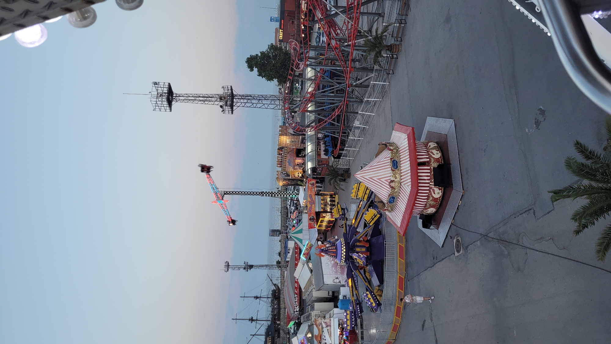 This image captures an amusement park scene, likely in Varna, Bulgaria, during twilight. The sky is a soft gradient of pale blue and grey, indicating either early morning or late evening. Various amusement rides are visible, including a roller coaster with red tracks, a tall drop tower with a checkered flag pole, and a spinning ride with purple and yellow elements. A carousel with a red and white striped canopy, adorned with decorative figures, is also prominent. In the background, a large sailing ship structure suggests a maritime theme within the park. Several people are present, some walking on the paved ground, and others appearing to be on or near the rides. The atmosphere seems relaxed, with the park appearing to be open for visitors. The lighting from the rides and surrounding structures is starting to become more noticeable as daylight fades.