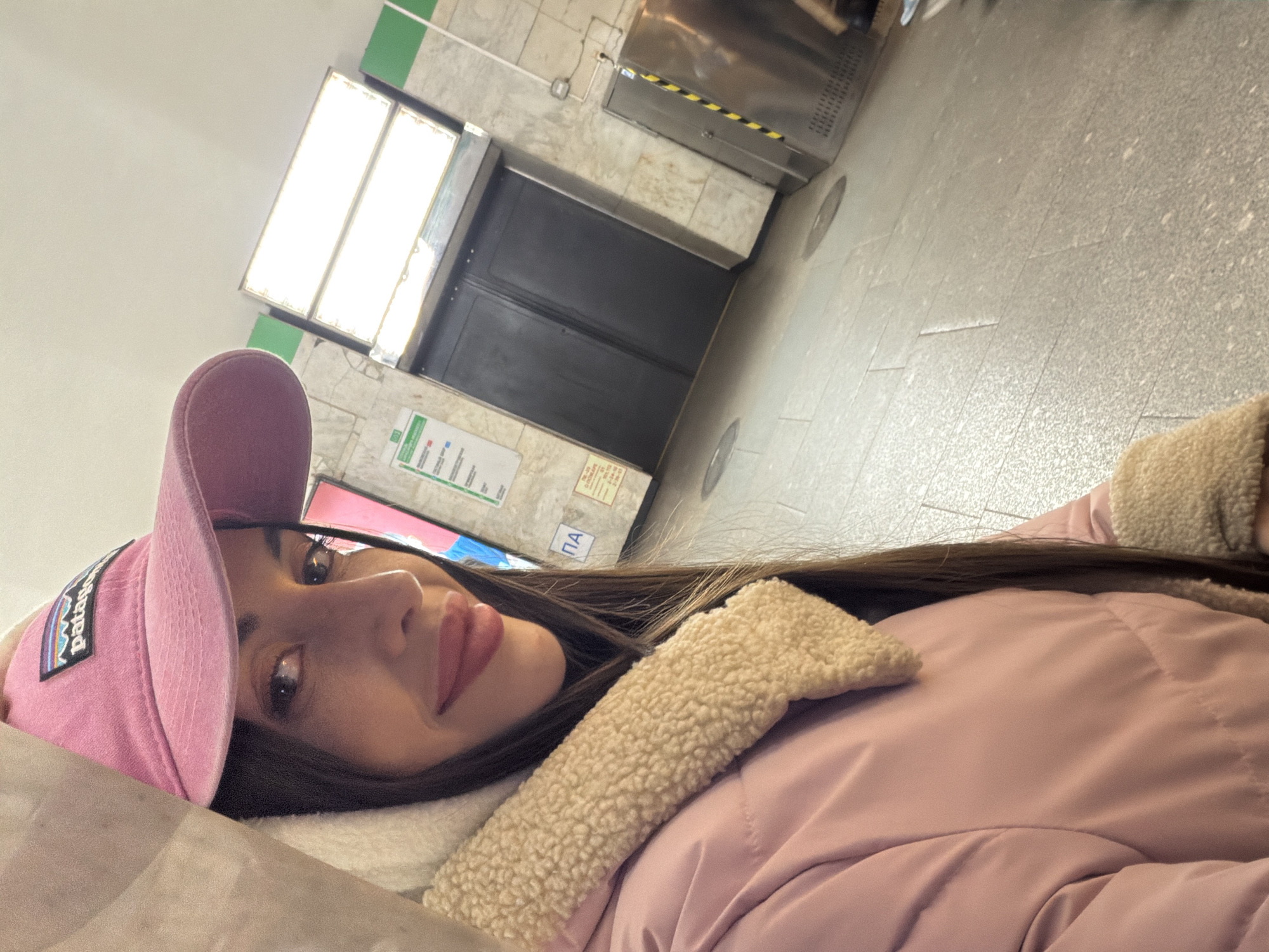 A young woman is lying down in what appears to be a subway station. She is wearing a pink jacket with a sherpa-style collar and a pink baseball cap with a visible Patagonia logo. The subway station has light-colored tiled walls and floors, and there is a service elevator in the background. A sign with "ПA" is visible on the wall. Based on the station's architecture, typography, and overall appearance, it is plausible this photo was taken in a subway station in Saint Petersburg, Russia.