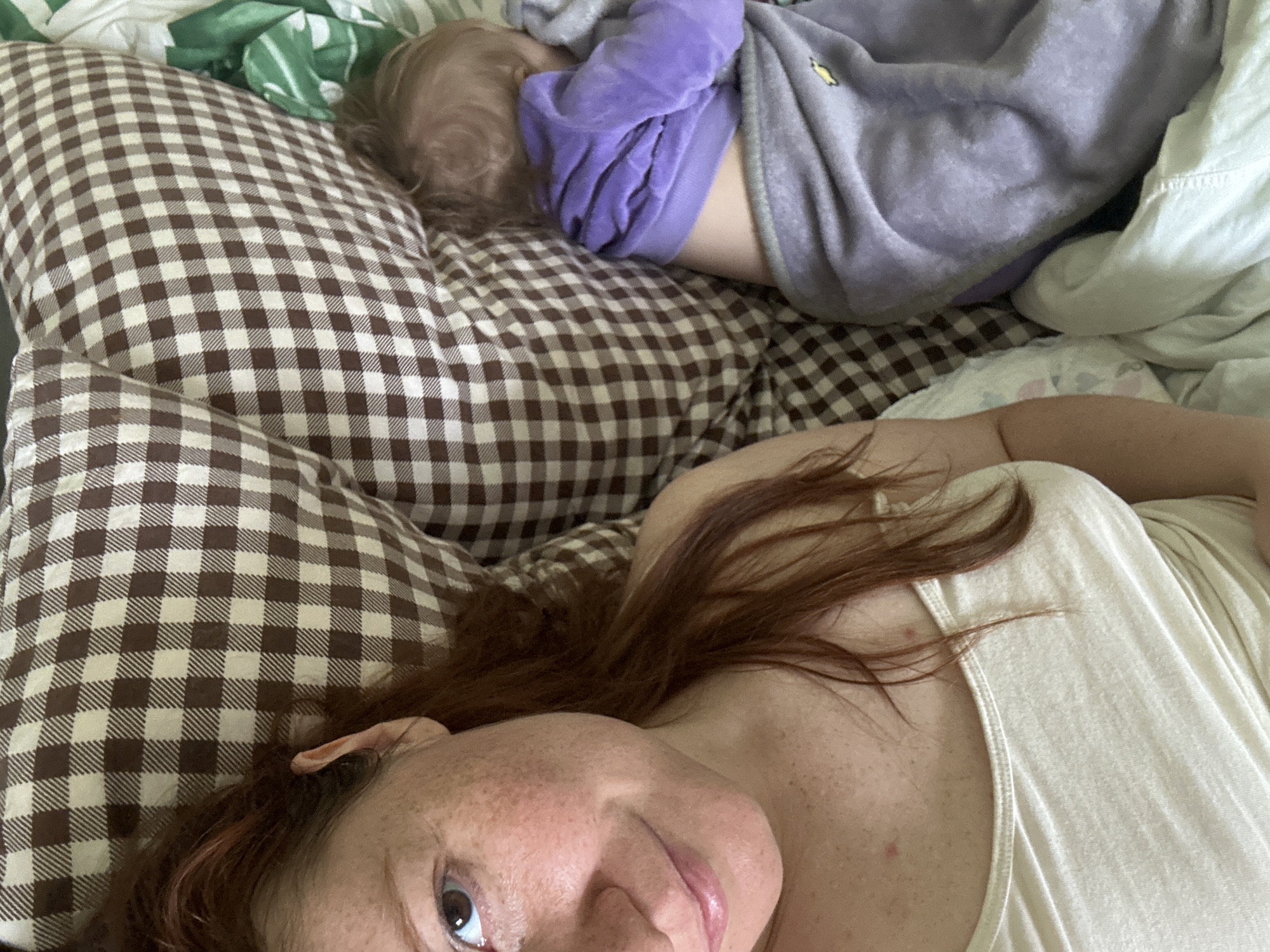 A woman lies on a bed with a brown and white checkered pillow. Next to her, a child is asleep, wearing a purple and gray outfit. The woman has red hair, freckles, and is wearing a light-colored tank top. The background includes a green patterned blanket. The location in the city of Nizhny Novgorod, Russia cannot be determined from this image.