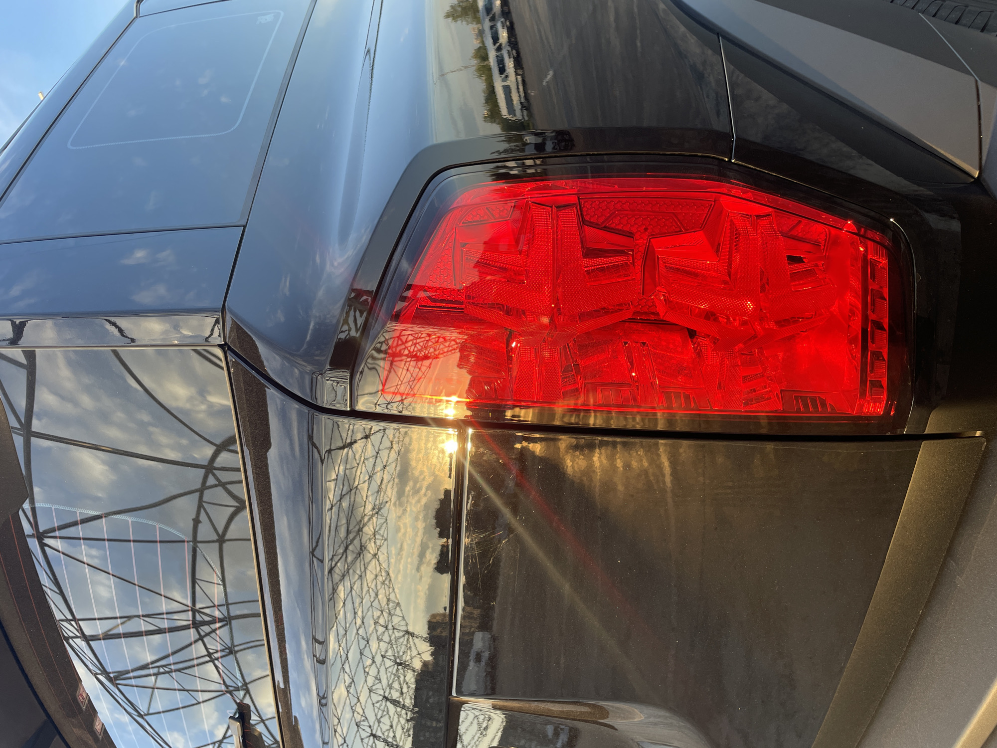 This media file is a close-up, eye-level, outdoor shot of the rear of a dark-colored car, likely taken in Kazan, Russia. The primary subject is the car's bright red taillight, which is illuminated and reflects the sun's glare. The taillight has a complex, geometric internal design with multiple facets and clear red lenses.
The car's body panels are dark and highly reflective, mirroring the sky, clouds, and a metal structure, possibly a geodesic dome or scaffolding, suggesting the car might be in a parking lot or an open area with such a structure nearby. The reflections on the car's surface indicate it is daytime, with bright sunlight, possibly late afternoon given the warm golden tones in the reflections. The sky appears clear with scattered clouds.
Notable details include what appears to be a panoramic sunroof or a tinted glass panel on the roof of the car, visible in the upper left portion of the image. There are also what look like minor scratches on the car's paintwork below the taillight, catching the sunlight. The textured, matte plastic trim of the bumper is also visible at the bottom. No people are visible in the image. The overall impression is a detailed view of a vehicle's rear, emphasizing its design and the interplay of light and reflection.