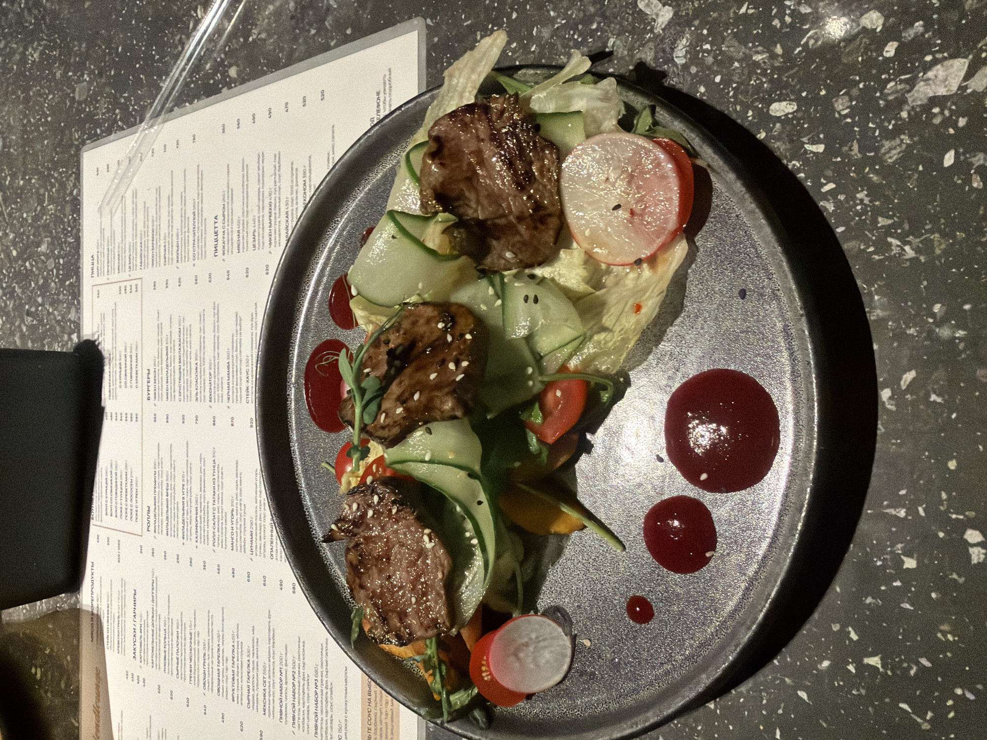 The image depicts a plate of food on a dark, speckled surface, presumably a table. The plate contains a salad with lettuce, cucumber ribbons, radish slices, tomato pieces, and what appears to be seared meat, possibly beef, sprinkled with sesame seeds. The food is garnished with red sauce spots. A menu is visible on the left, with text in Russian. The image appears to be taken from an overhead perspective. Given the Russian text on the menu, it is likely the location is in Russia. I don't have the information to confirm whether the image was taken in the city of Chaikovsky, Russia.