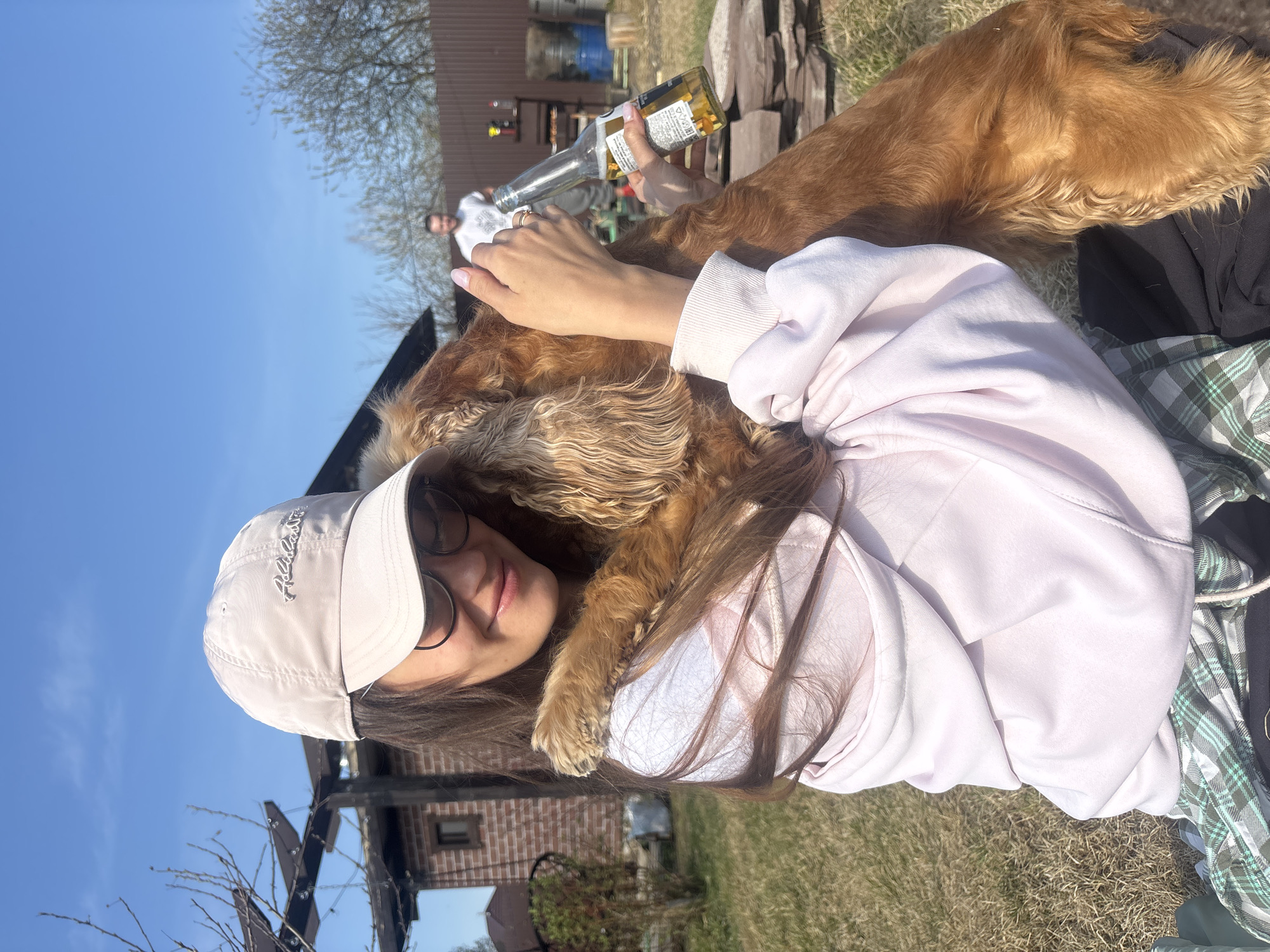 A woman wearing a light pink hoodie and a light-colored baseball cap is holding a brown dog in her arms. She is wearing glasses and has long brown hair. The dog is resting its paw on her shoulder. In the background, there is a brick building, a wooden structure, and a person in a white shirt. Someone is holding a beer bottle behind the dog. The location is a grassy area, possibly outdoors. The city is Emel'yanovo, Russia.