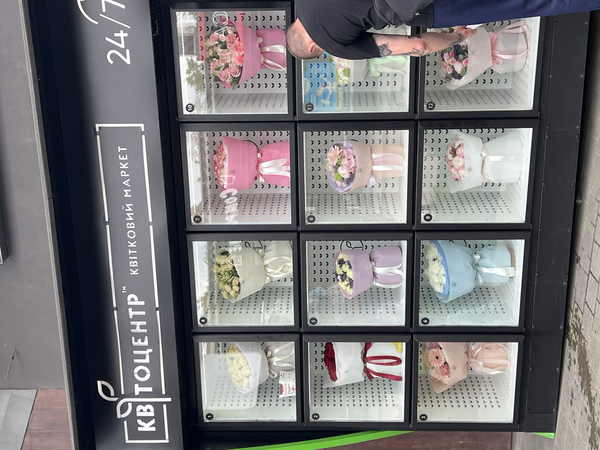 The image depicts a flower vending machine in Ternopil, Ukraine. The machine is black and rectangular, with twelve individual glass-fronted compartments displaying various bouquets of flowers wrapped in paper and ribbons. Each compartment is numbered. A man, partially visible in the upper right, is selecting flowers from the machine. He has short hair, a black shirt, and tattoos on his arm. The vending machine has "KB CENTER" and "KVITKOVYI MARKET" written vertically on the side. The background is blurred, suggesting an urban environment. The machine also has “24/7” written on the top, indicating that it operates around the clock.