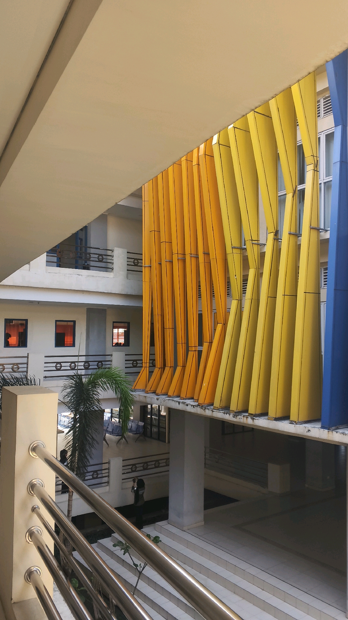 The image captures a view of an architectural structure with a prominent facade featuring alternating vertical elements in shades of orange and yellow, along with a section in blue. These elements are angled and form a visually striking design. The building appears to be a multi-story structure, with balconies and windows visible on the white-colored floors. A palm tree is situated to the left of the orange and yellow facade. In the foreground, a stainless steel railing is visible, suggesting a walkway or balcony. The scene is set within an open courtyard or atrium, with stairs and seating areas visible in the lower portion of the image. Two individuals are present in the lower section; one is standing near a tree and the other is further back near the stairs. The overall impression is that of a modern public building or educational institution. Based on available information, this architectural feature is located at the STIKOM Poltekgo in Lohbener, Indramayu Regency, West Java, Indonesia, which is part of the broader Lohbener area.