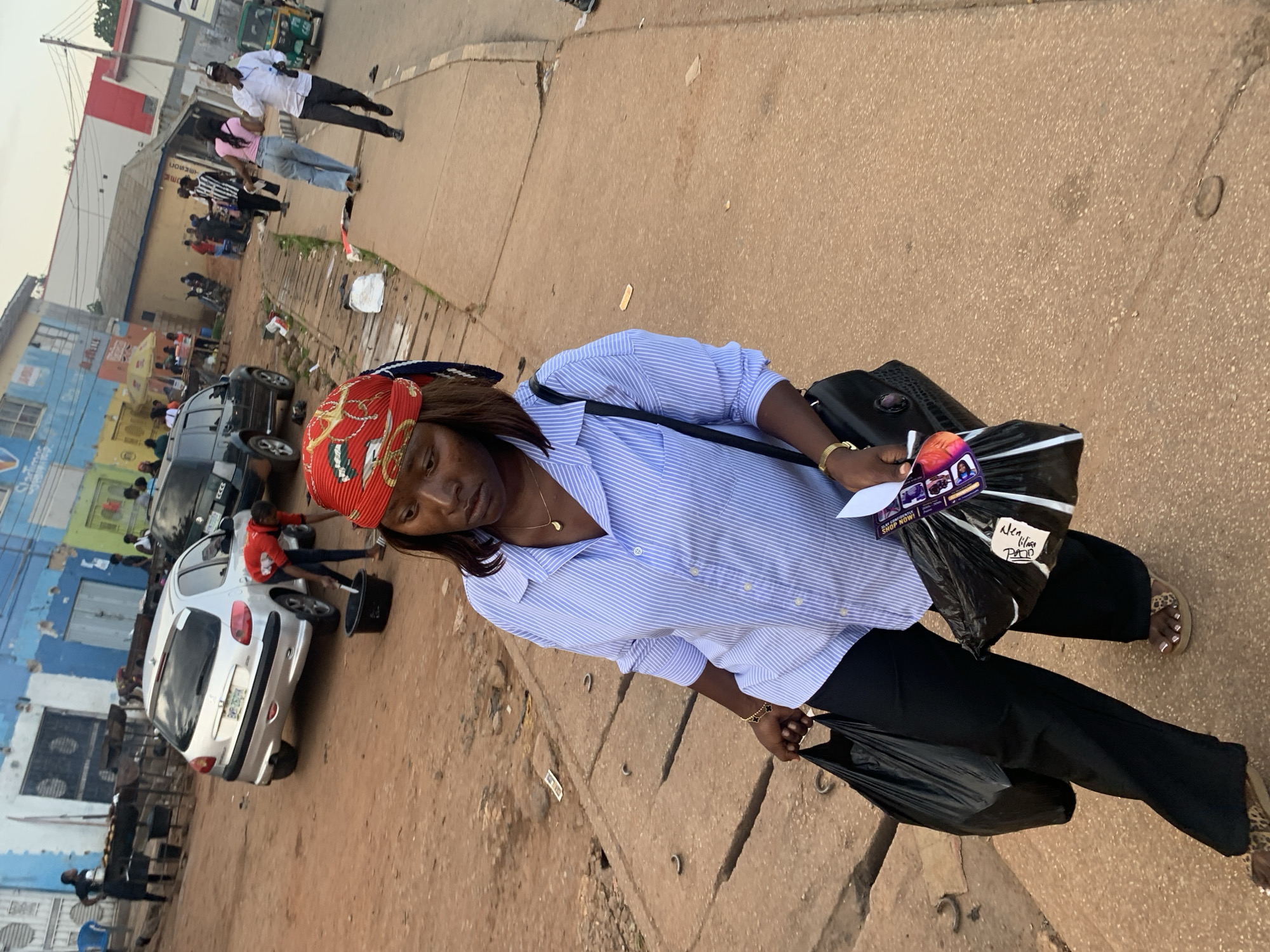 The image captures a street scene in Barakin Lerawi, Nigeria, under an overcast sky. In the foreground, a woman walks along a paved sidewalk, carrying two black plastic bags and a small purple and white package. She is wearing a blue and white striped button-down shirt, black pants, and leopard-print sandals. A gold bracelet adorns her left wrist, and she has a red and gold patterned headscarf. Behind her, a dirt road is visible, with several vehicles including a silver Peugeot 206, a dark SUV, and a green auto-rickshaw. In the background, buildings with colorful facades line the street, and several people are seen in various activities, some appearing to be vendors at stalls. The environment appears to be a bustling marketplace or commercial area.
